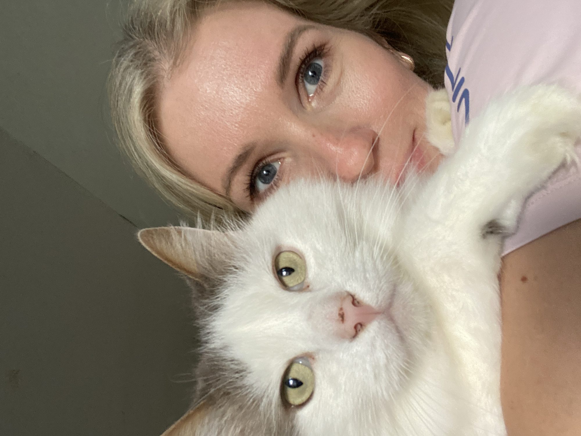 A close-up, selfie-style photograph depicts a fair-skinned woman with blonde hair and blue eyes holding a predominantly white cat with olive-green eyes. The woman is angled towards the upper portion of the frame, with her face slightly tilted. She wears a light pink garment that is subtly visible. The cat occupies the lower portion of the image, its face and upper body prominently displayed. The cat's nose is pink with small dark spots. The background is blurred and indistinct, ranging from light to dark. The location appears to be indoors, with no specific city or identifying details available.