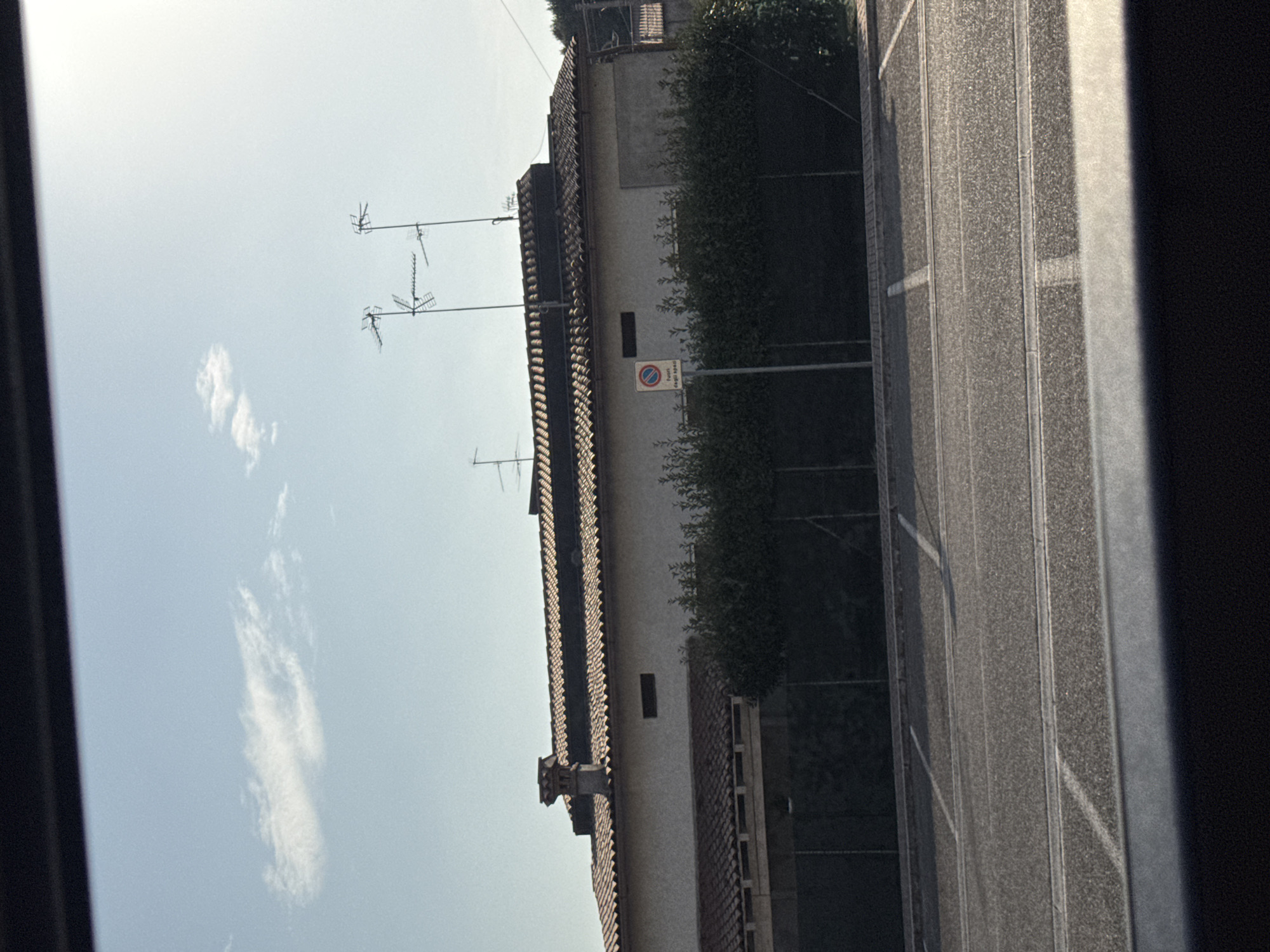 The image captures a view of a building with a terracotta tiled roof and a chimney, situated behind a hedge and a fence. Two sets of television antennas are visible on top of the building. In the foreground, a paved area with white parking lines is present. The sky is clear with a few clouds. A circular sign with red and blue markings and text in Italian is mounted on a pole in front of the hedge. The scene appears to be located in an outdoor urban or suburban environment, possibly a parking lot adjacent to a residential or commercial building.