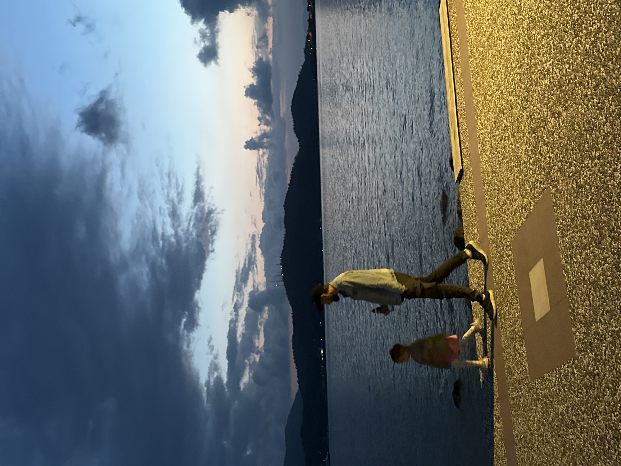 The image captures an evening scene at dusk along a brightly illuminated waterfront promenade in Fethiye, Turkey. Two individuals, an adult male and a child, are depicted walking from right to left across the foreground. The adult male, silhouetted against the water, wears a dark cap, a light-colored, possibly denim-style jacket, dark trousers, and dark athletic shoes with light soles; he carries a dark bottle in his right hand. Slightly ahead of him is a child, also partially silhouetted, wearing a light-colored top, a pink skirt or dress, light leggings, and light shoes.
The immediate foreground features a textured, light-colored aggregate promenade surface, strongly illuminated by an unseen artificial light source to the right, creating warm tones. This surface incorporates darker, reddish-brown rectangular and linear decorative patterns. Adjacent to the promenade, a calm body of water, likely the Fethiye Bay, extends into the mid-ground, with faint ripples visible. A dark wooden plank or similar structure rests at the water's edge on the far right.
Across the water, dark, undulating hills or mountains are silhouetted against a sky transitioning from a deep twilight blue near the horizon to darker, heavy grey clouds overhead. Sparse artificial lights are visible on the distant hillsides, indicating structures or distant settlements. The overall atmosphere is one of peaceful evening activity along a well-lit coastal path.