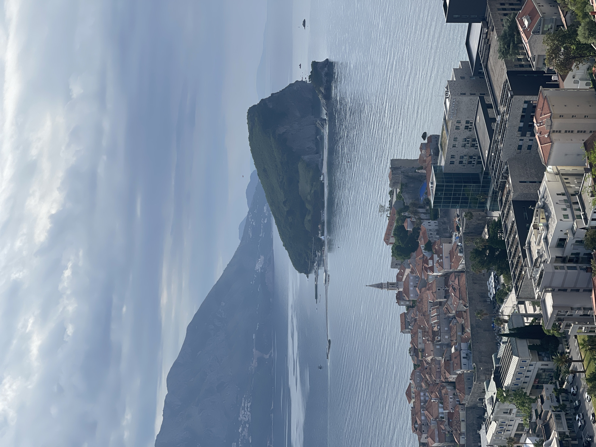 The image captures a panoramic view in Budva, Montenegro. The Adriatic Sea dominates the view, with a rocky islet covered in vegetation positioned prominently. To the left is a mountain range partially obscured by haze, and the coastline runs from the mountains towards the city. The city of Budva is situated on the right side of the image, with a mix of old town buildings featuring red-tiled roofs and modern structures. A distinct church steeple rises above the old town buildings. The sky is overcast, with clouds covering a portion of the horizon. The image is taken from an elevated position, offering a broad view of the landscape.