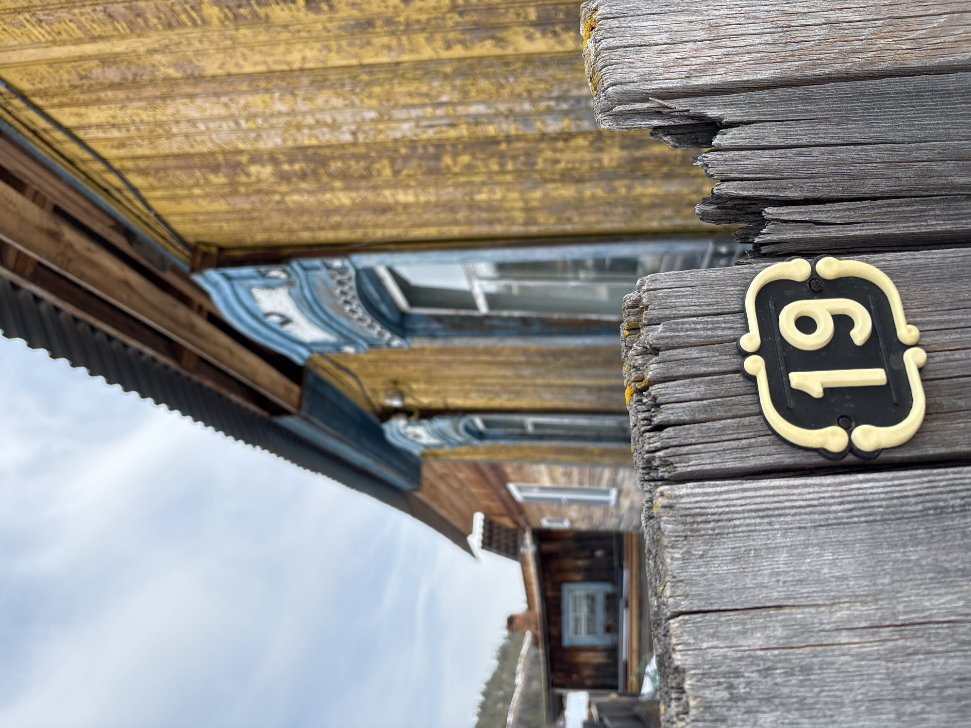 A weathered wooden house in Невон, Russia, features the house number "61" on its exterior. The gray planks of the house wall are slightly weathered, with some signs of wear and tear. The number "61" is displayed on a small, decorative plaque with a black background and light-colored numbers. The house itself is painted in shades of yellow and blue, with some visible wood grain. The sky is overcast.