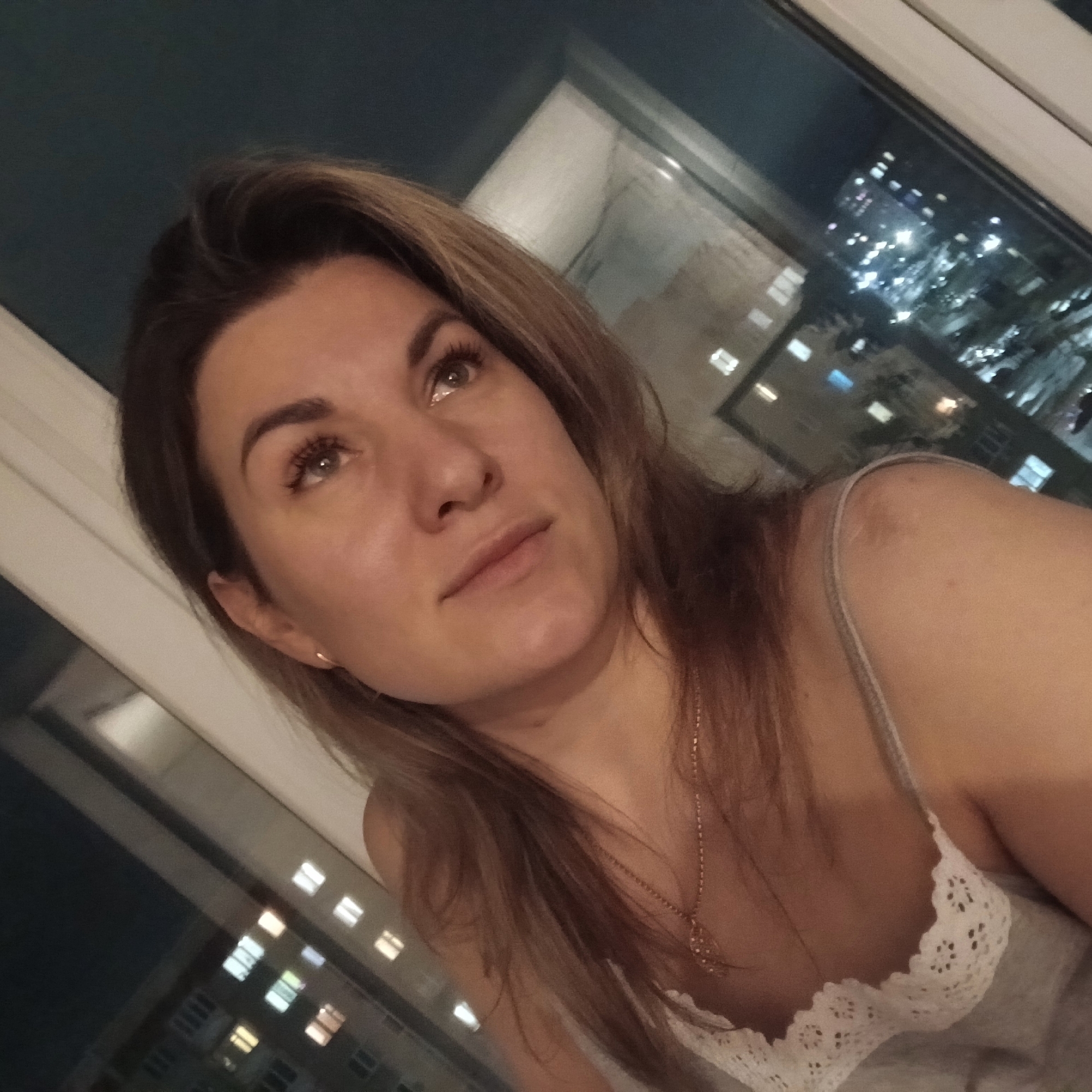 A woman with fair skin and medium-length brown hair with blonde highlights poses indoors near a window. She is wearing a light-colored, possibly white, tank top with lace detailing. A gold necklace hangs around her neck. Her gaze is directed upwards and slightly to the left. Outside the window, a nighttime cityscape is visible with illuminated buildings, suggesting an urban setting. Based on the appearance of the buildings in the background the location may be in the city of Izhevsk, Russia.