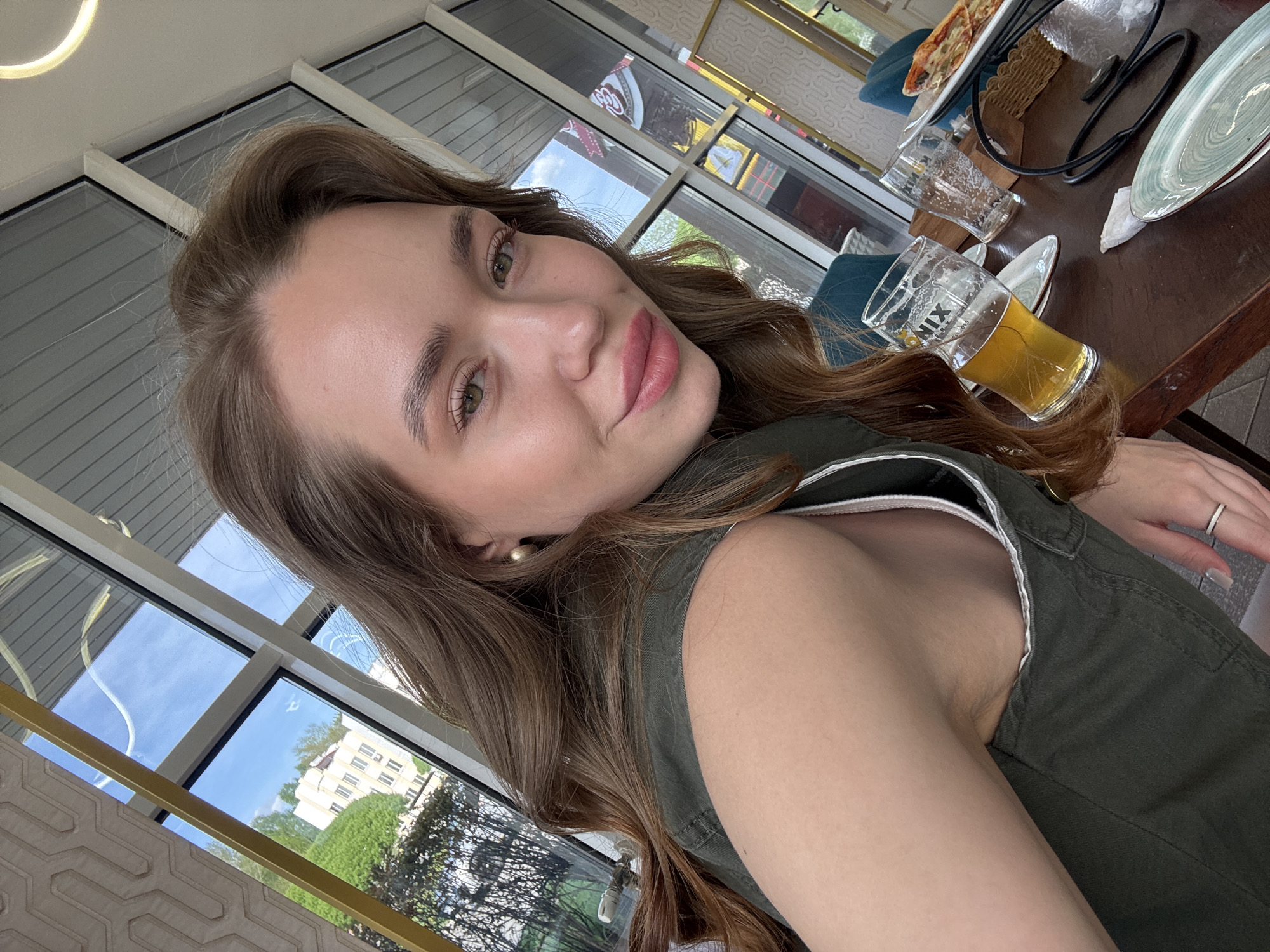 A young woman poses for a selfie indoors near a window. She has long brown hair and wears a dark green sleeveless top. A partial view of a table reveals a glass of beer, a plate with leftover food, a pair of eyeglasses, and stacked plates. The background shows a building with trees outside. The image was taken in the city of Chaikovsky, Russia.