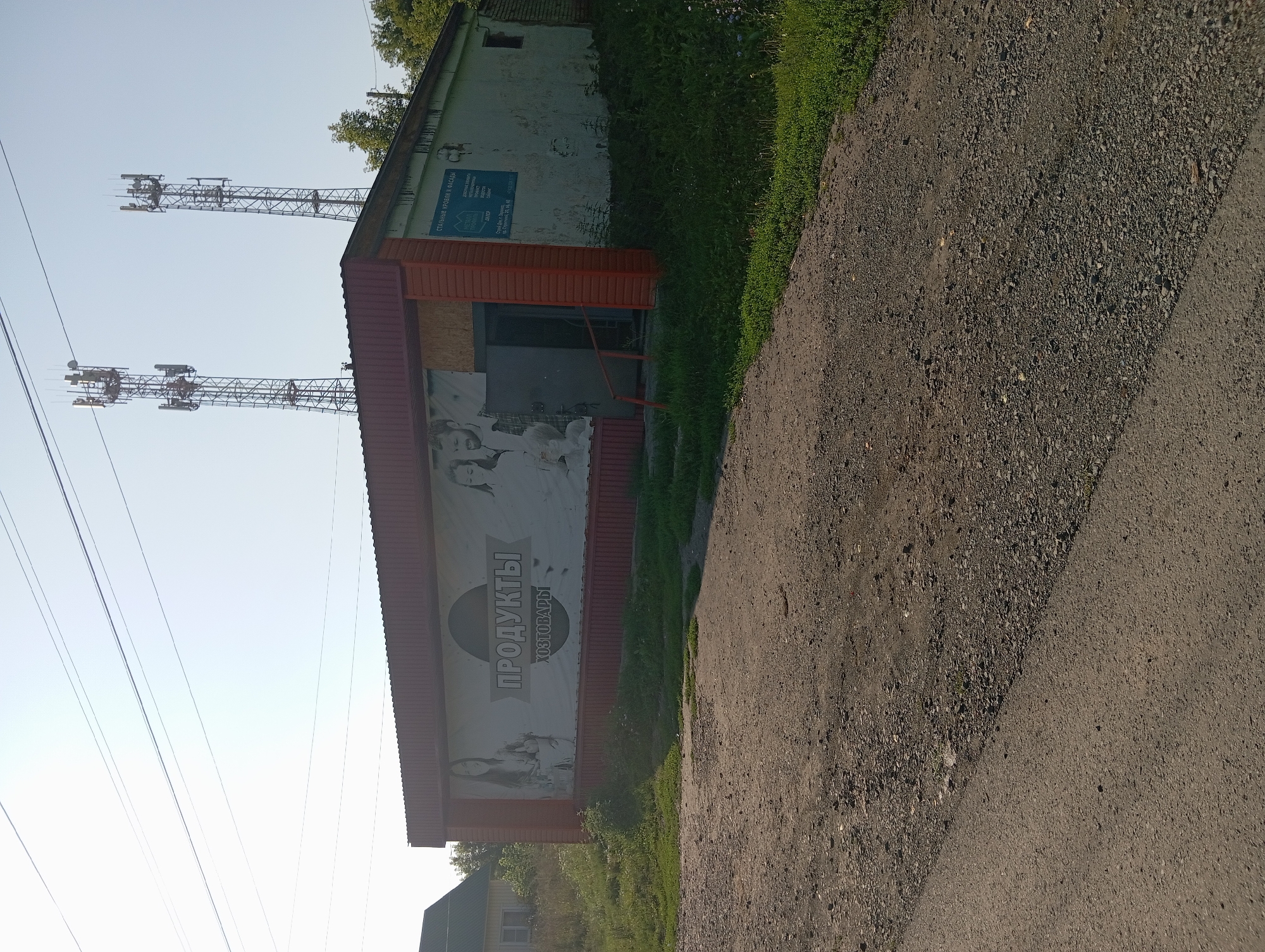 The image shows a small, single-story, rectangular building with a red corrugated metal roof. A large banner on the front of the building displays the word "ПРОДУКТЫ" (Products) in large, stylized letters, along with the word "ХОЗТОВАРЫ" (Household goods) below it, and images of people. To the right of the banner, on the building's facade, is a blue sign with text and logos, including "СТАЛЬНЫЕ КРОВЛИ И ФАСАДЫ" (Steel roofs and facades). The building has a doorway with steps leading up to it and a railing. The surrounding environment includes green grass, some trees, and a dirt and gravel road in the foreground. In the background, two tall telecommunications towers are visible against a clear blue sky. A residential house with a green roof is partially visible to the left of the main building. The image appears to be taken in NovoKopylovo, Russia.