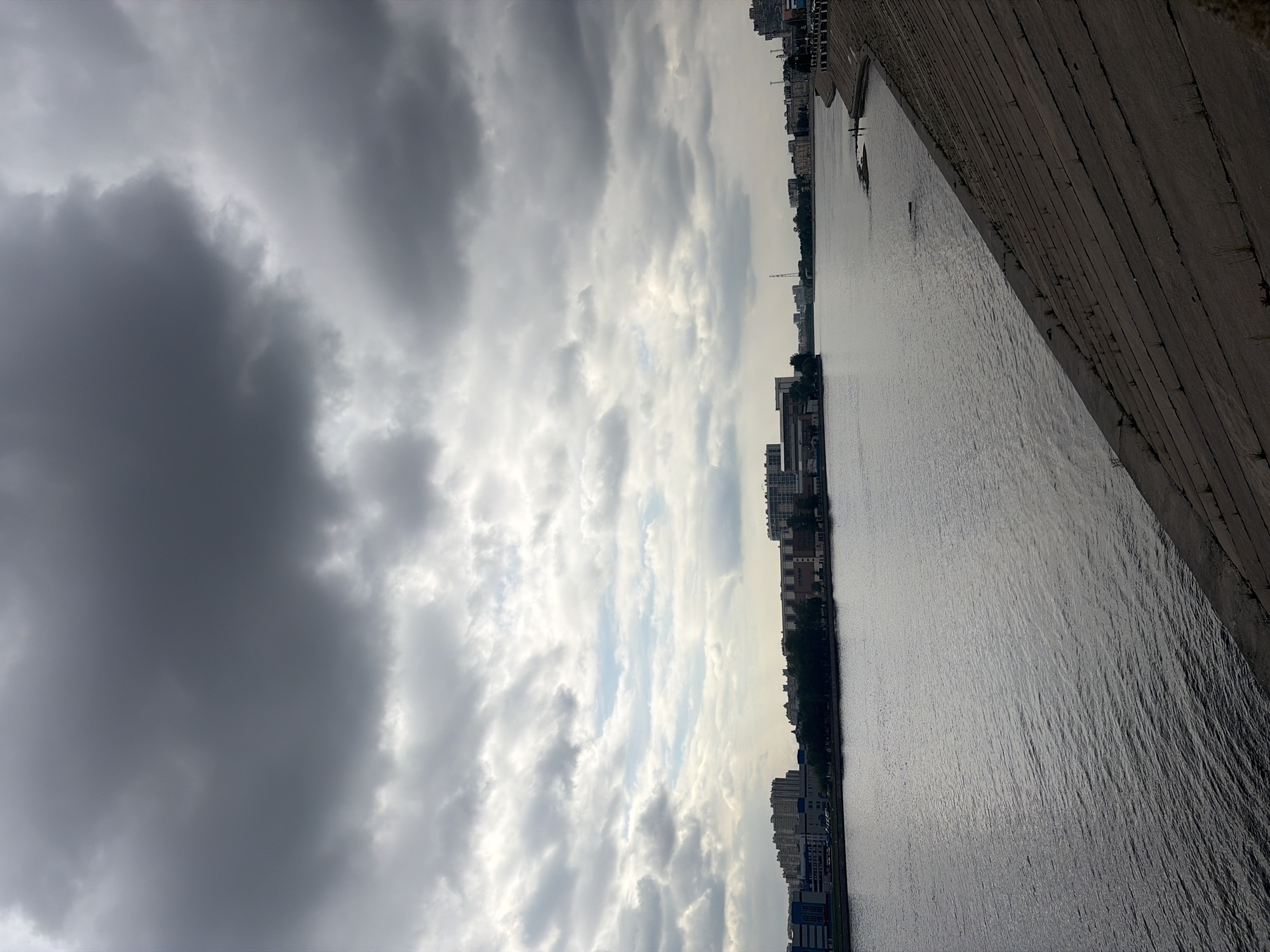 The image depicts a waterfront scene in Saint Petersburg, Russia. The view is taken from an elevated position, likely a bridge or embankment, looking out over a body of water, possibly the Neva River or a canal. The water surface appears calm with subtle ripples.
On the right side of the frame is a concrete embankment structure, angling upwards and out of view. On the opposite bank, a line of buildings and trees forms the city skyline. The architecture suggests a mix of residential and commercial structures.
The sky occupies a significant portion of the image, filled with a variety of cloud formations. Dark, dense clouds dominate the left side, contrasting with lighter, partially illuminated areas towards the center. The overall atmosphere is overcast, creating a subdued lighting.
There are no people visible in the image. There are a few small boats visible on the water.