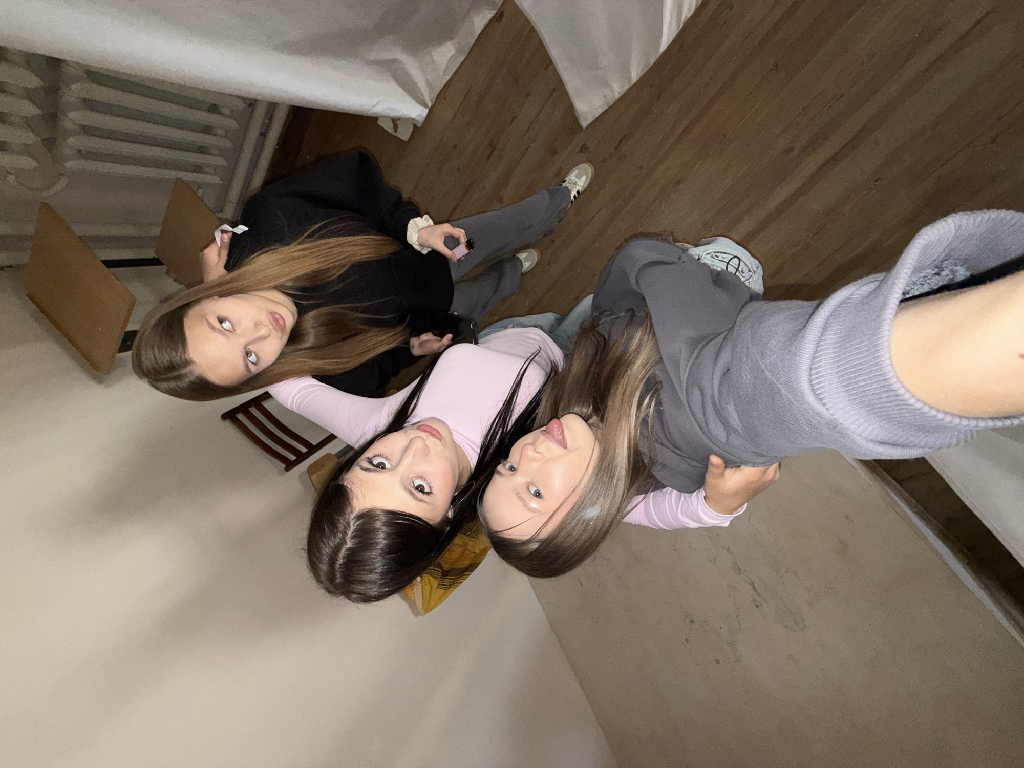This high-angle, tilted image captures a group selfie of three young women indoors. The perspective suggests the photo was taken from above with an extended arm or camera.
The woman positioned on the left side of the frame has long, straight brown hair and is wearing a black top or hoodie paired with grey pants and white sneakers. She gazes directly at the camera, holding a small pink object, possibly a lighter or vape, in her hand.
In the center, another woman with long, dark brown hair wears a light pink long-sleeved top. She looks towards the camera with a playful expression, her tongue slightly extended.
The third woman, closest to the camera on the right, has long, light brown hair and is wearing a grey sweatshirt over a light pink and white striped long-sleeved top. Her arm is visible, indicating she is likely holding the camera. Her gaze is also fixed on the lens.
The setting appears to be an indoor room featuring warm brown wooden floorboards and light-colored walls or ceiling. A radiator and parts of wooden chairs or stools are discernible in the background. The lighting is consistent with an indoor environment. The subjects convey a relaxed and friendly demeanor. There is no visible text in the image. While the location context of Chaikovsky, Russia is provided, no specific visual cues within the image confirm this.