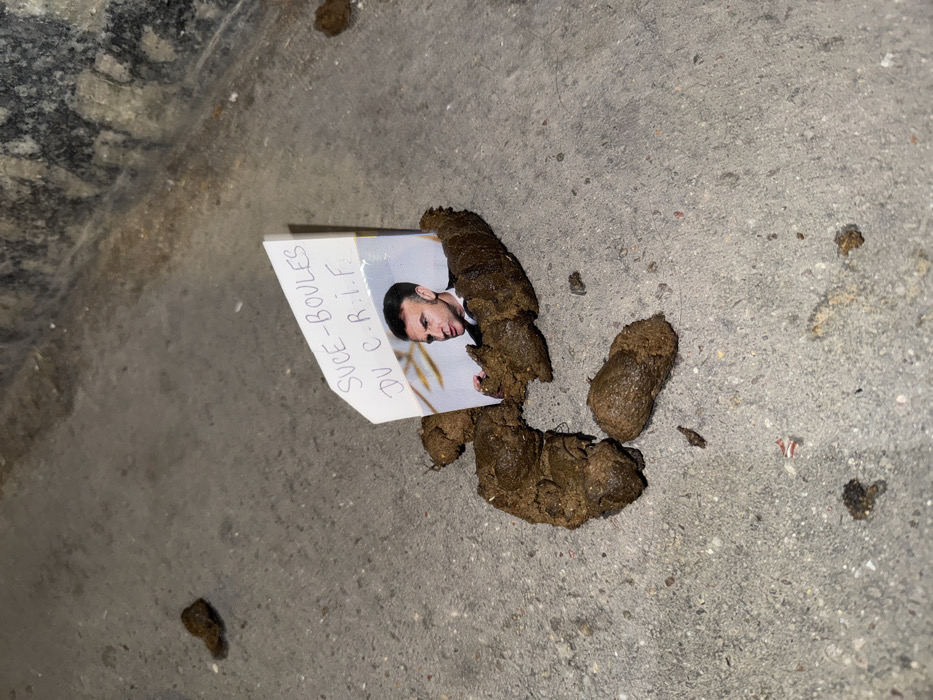 The scene depicts a deliberate arrangement of animal feces on a light gray, textured concrete or asphalt ground. A small, white piece of paper, folded to stand upright, is embedded within a central mass of dark brown, semi-solid fecal matter. This paper features a color photograph of Emmanuel Macron, President of France, wearing a dark suit and tie. Above the photograph, handwritten in black capital letters, is the phrase "SUCE-BOULES DU C.R.I.F." Several smaller, individual pieces of feces are scattered around the main arrangement. The ground surface shows minor discolorations and small fragments of debris. In the upper right background, a darker, possibly stained and textured concrete or stone surface, consistent with a wall or curb, is visible. The exact location within the city of Paris, France, is not visually discernible from the image, but the identifiable individual and French text indicate a context within France.