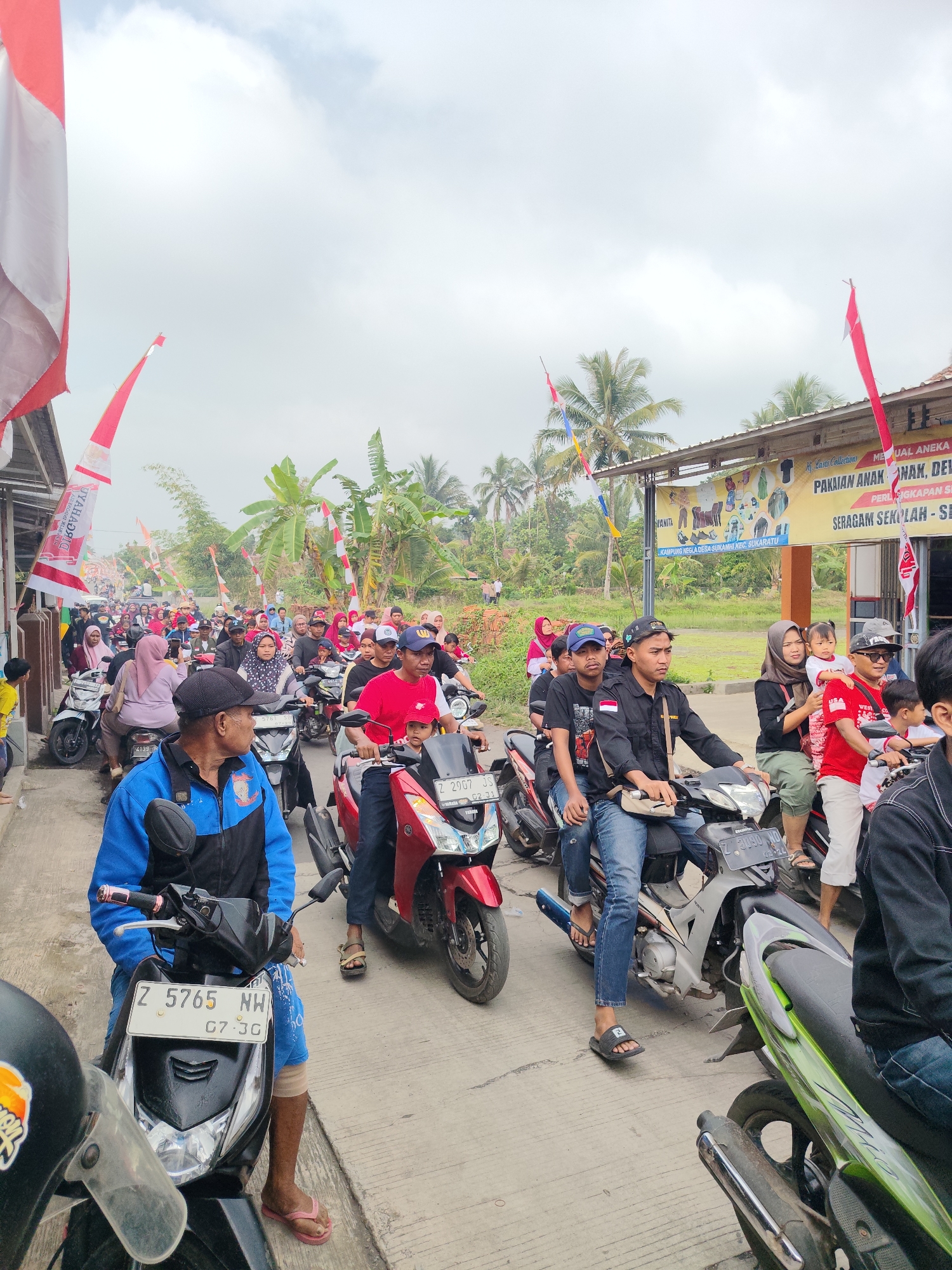 The image captures a lively street scene in Sukaratu, Indonesia, on an overcast day. A convoy of motorcycles fills the road, with many people riding on them. The atmosphere appears festive, indicated by numerous red and white flags and banners adorning the street and buildings. One banner, displayed on a shopfront, reads "MENJUAL ANEKA PAKAIAN ANAK, DEWASA, SERAGAM SEKOLAH - SEPATU". The people are dressed in casual attire, with some wearing red and white, possibly in celebration of a national holiday. The motorcycles are predominantly modern scooters, with a few older models visible. The road surface is asphalt, and the buildings lining the street are simple structures with tiled roofs. Lush green vegetation, including palm trees and banana trees, can be seen in the background, suggesting a rural or semi-urban setting within Sukaratu. The people appear engaged and are looking in various directions, some towards the camera, others ahead. The overall impression is one of community gathering and movement, possibly for a parade or celebration.