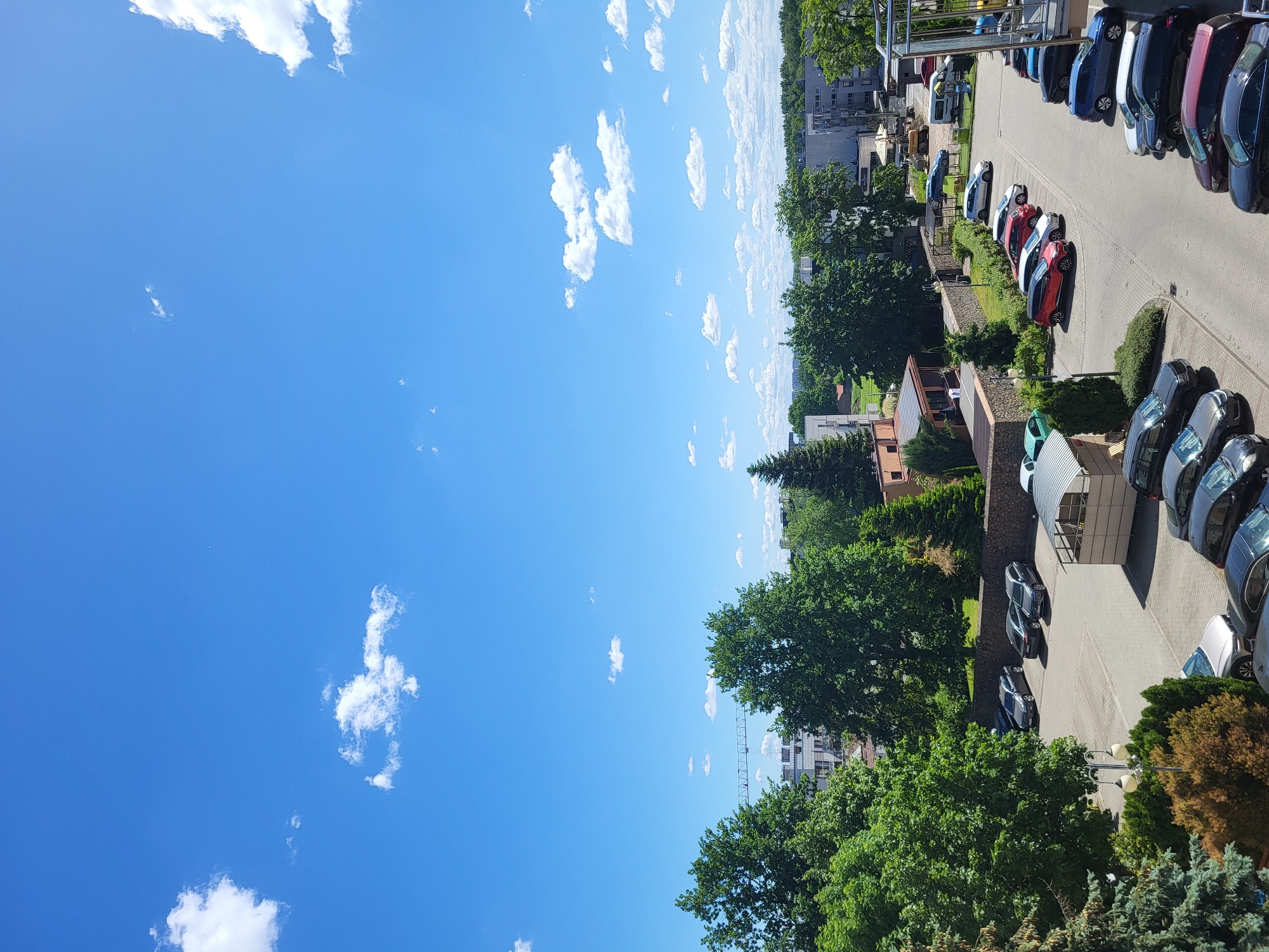 Here is a matter-of-fact description of the image:
The image presents an elevated view of an outdoor parking lot adjacent to a cluster of buildings, possibly a residential or commercial complex. The sky occupies a substantial portion of the left side of the image, displaying scattered cumulus clouds against a blue backdrop. The right side is dominated by a parking area where several cars are aligned. Vegetation, primarily trees and shrubs, is visible between the parking area and the buildings. The buildings appear to be a mix of residential and commercial structures. The general lighting suggests it is daytime, likely midday.