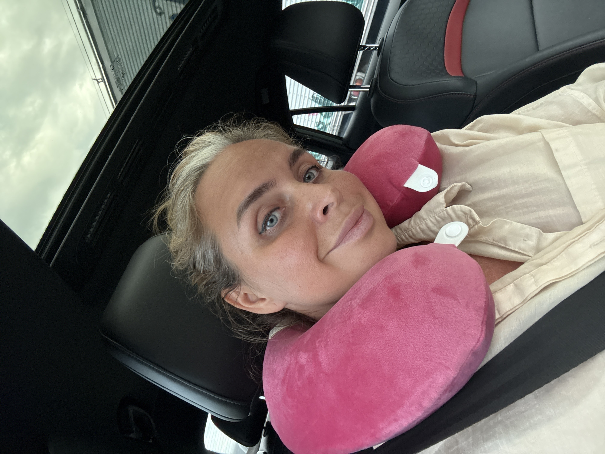 A woman with blonde hair and blue eyes, wearing a cream-colored shirt and a pink neck pillow, is sitting in a car with a sunroof. She is looking at the camera and smiling. The interior of the car is visible, including black leather seats with red accents and a black seatbelt. The image appears to be taken during the daytime. There is no indication of the specific location within the city of Ban Nong Bua, Thailand.