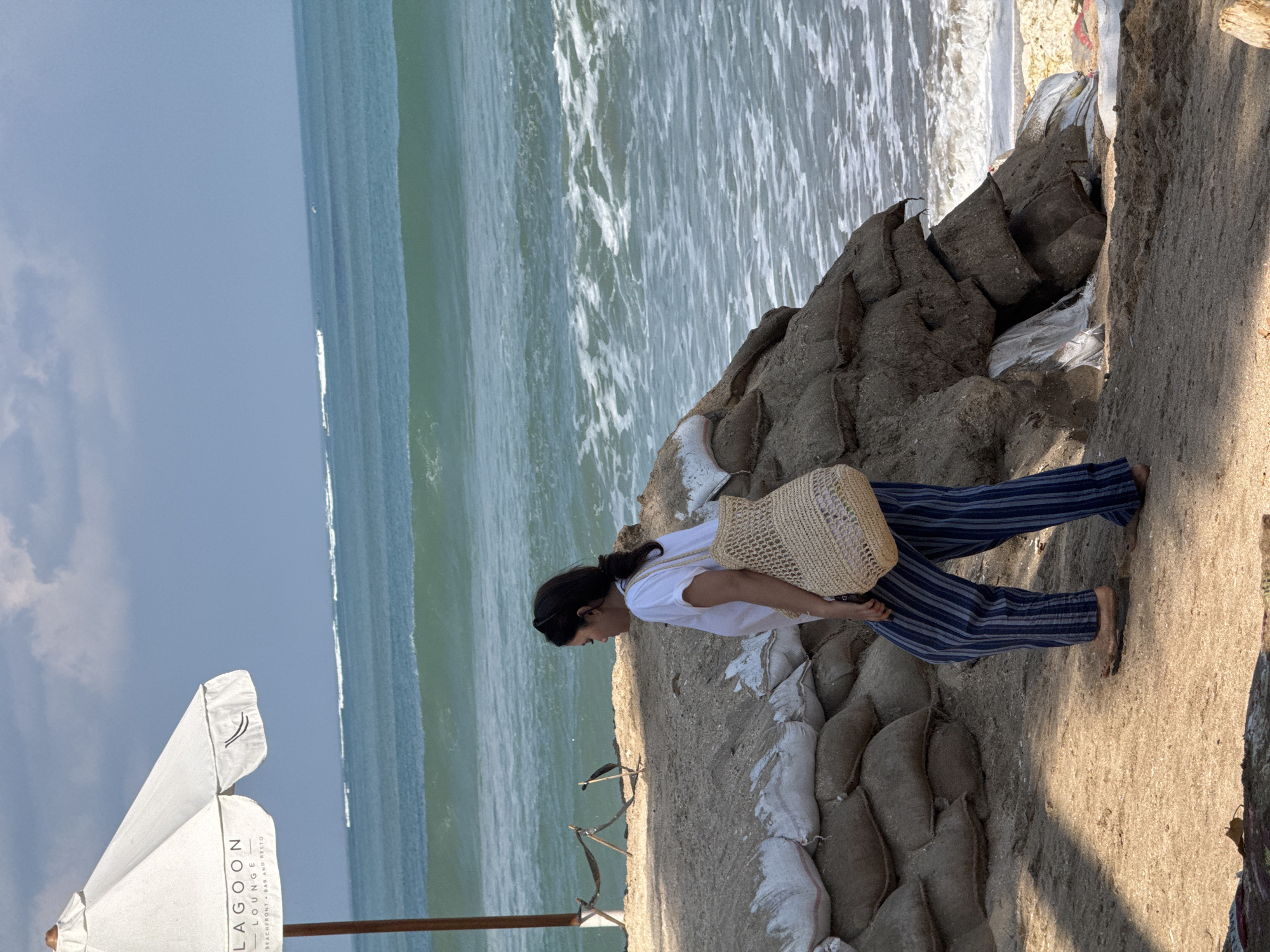 A woman walks along a sandy beach in Kuta, Indonesia, near sandbag barriers and a white umbrella with the text "Lagoon Lounge Beachfront Bar and Resto". The woman is wearing a white t-shirt, blue and white striped pants, and carrying a woven bag. The ocean is visible in the background with gentle waves breaking on the shore. The sky is partly cloudy.