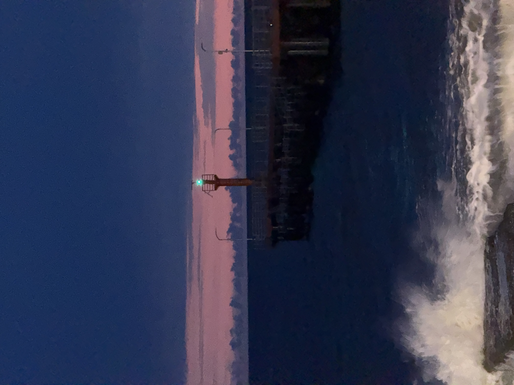 Here is a description of the image:
The image depicts a seaside scene, likely a harbor or port, with a prominent lighthouse emitting a green light. The water is dark blue, with white foamy waves crashing against a structure, possibly a pier or breakwater. A series of streetlights line the area near the water's edge. The sky transitions from a deep blue to a pinkish hue, suggesting either dusk or dawn. The overall atmosphere is tranquil and serene. The location is likely a coastal city like Sochi, Russia.