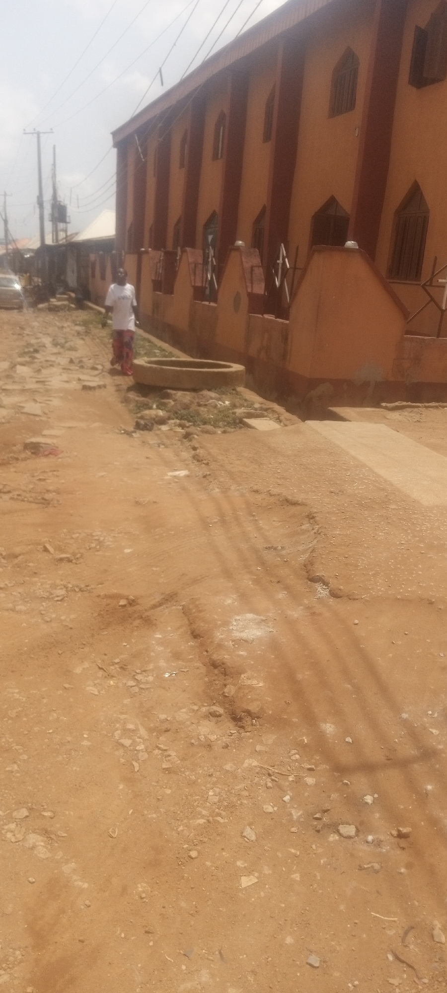 The image depicts a dirt road in Narayi, Nigeria. The road is uneven and has potholes. A man is walking on the side of the road near a concrete well. The building to the right is painted orange and has brown trim. There is a car parked on the left side of the road and power lines overhead. The sky is hazy.