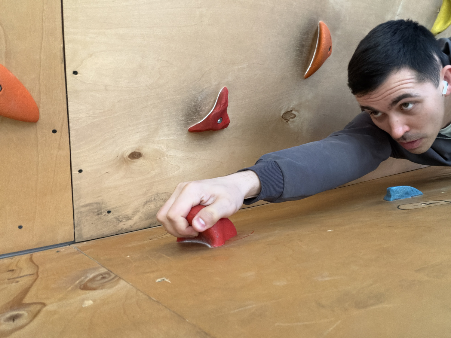 An eye-level shot captures a young adult male actively engaged in indoor rock climbing or bouldering in Izhevsk, Russia. The man, likely in his 20s or 30s, has short dark hair, dark eyebrows, and a focused, determined expression. He wears a dark grey long-sleeved sweatshirt and a white wireless earbud in his right ear. His right hand is firmly gripping a bright red climbing hold on a light-colored plywood panel that forms part of the climbing wall.
The climbing wall is constructed from multiple light-colored wooden panels, some vertical and others forming a horizontal surface or overhang section, all featuring visible wood grain and small black screw or bolt holes. Several other colorful climbing holds are visible: two orange ones on the vertical panel to the left and upper-middle, another red hold on the upper right vertical panel, and a small, low-profile blue hold on the horizontal panel to the right of the man's arm. The man's body is positioned low to the ground, with his gaze directed upwards and to the right, indicating concentration on his next move. The scene is brightly lit, typical of an indoor sports facility. No text is visible in the image.