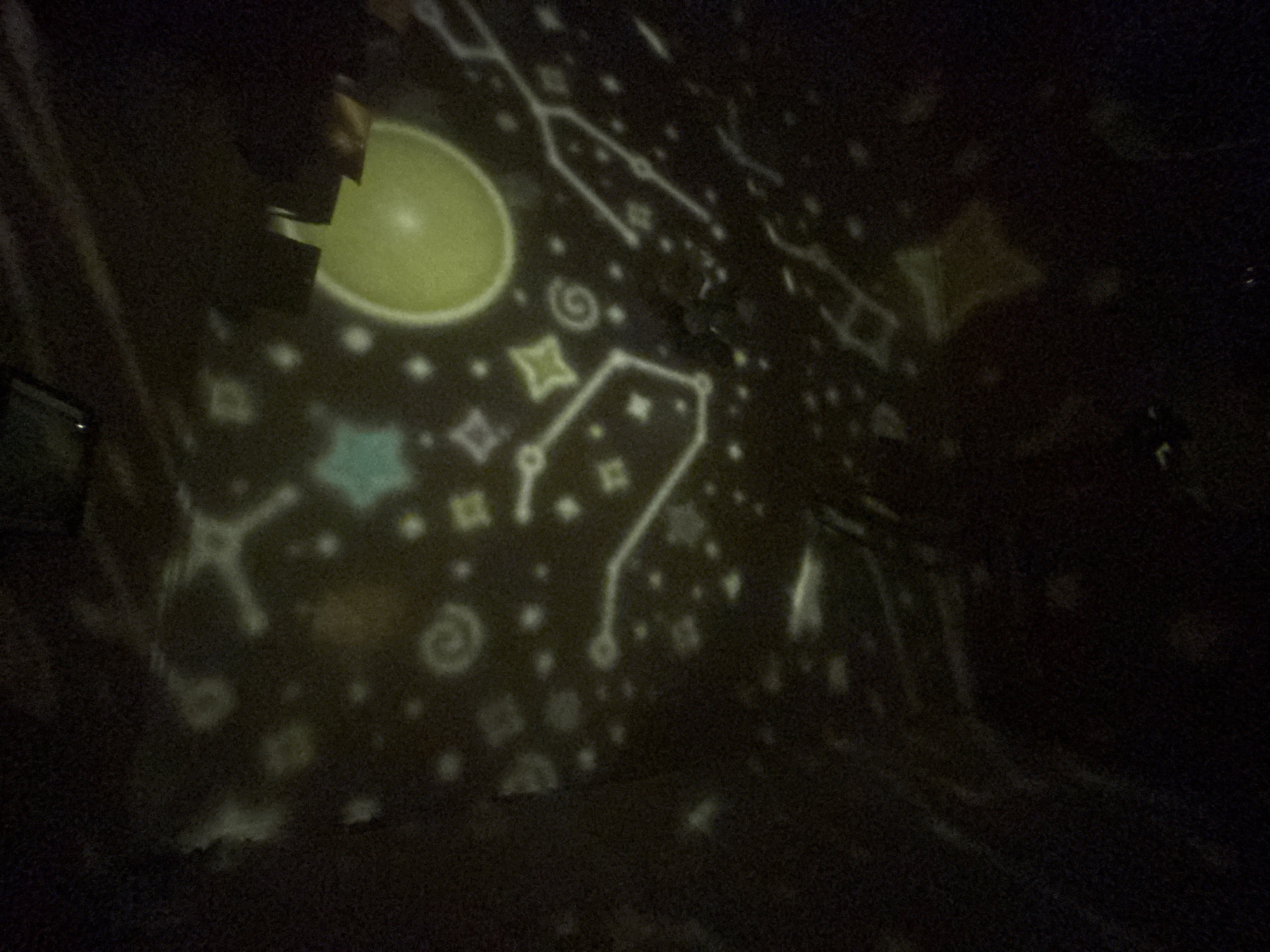 A projection of stars and celestial bodies is cast onto a dark surface. Some of the projected shapes are outlines of constellations, while others are dots representing stars. There are also a few larger shapes, one of which appears to be a bright yellow disc. The projection is blurry and has a grainy texture. The scene is dark overall, suggesting it is nighttime or an indoor setting with the lights off. The crops show closer views of specific projected shapes and a hint of a ceiling or upper surface, with the overall impression being a room illuminated by a star projector. There are no identifiable people, specific objects, or actions taking place that would allow for a definitive location in Kryvyi Rih, Ukraine.