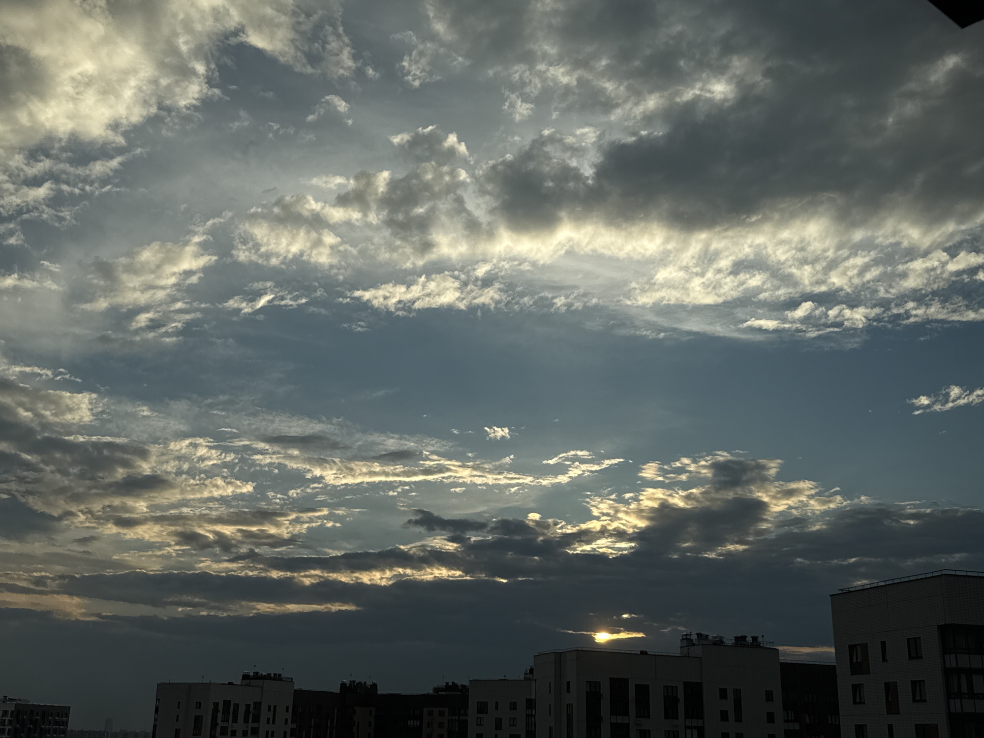 This media file is a photograph of a cloudy sky over a cityscape. The sky is filled with a variety of clouds, ranging from dark, heavy storm clouds to lighter, wispy formations. Sunlight filters through the clouds in some areas, creating a dramatic effect of light and shadow. The overall mood is somewhat somber and dramatic, with a sense of impending weather change.
In the foreground, the tops of several modern buildings are visible. These buildings appear to be apartment complexes, characterized by their multi-story structures and rectangular windows. The architecture suggests an urban or suburban setting. The buildings are silhouetted against the sky, with some details of their facades visible. The sun is setting or rising, as evidenced by the low angle of the sunlight and the warm tones in some parts of the sky. The time of day is likely early morning or late evening.
The location is identified as Lyubertsy, Russia. There is no visible text within the image. The scene captures a typical urban sky, with the interplay of natural elements like clouds and sunlight against the backdrop of man-made structures. The focus is on the sky and the atmospheric conditions, with the buildings serving as context for the scene.