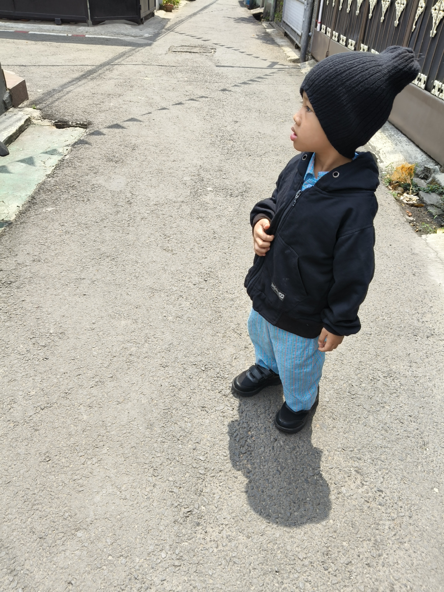 A young boy wearing a black beanie and a black zip-up hoodie over a blue checkered shirt stands on an asphalt road. He is also wearing blue checkered pants and black shoes with velcro straps. The boy is looking upwards and to the right. His shadow is cast on the road to his left. The road is a slightly worn asphalt surface, typical of residential streets. In the background, to the right, is a dark metal fence with decorative elements and some dry leaves and a patch of dirt on the ground next to it. To the left of the boy, the road edges with a concrete curb and a lighter green painted surface. The environment suggests an outdoor urban or suburban setting, potentially a street or alleyway in Bandung City, Indonesia, given the context. The lighting indicates a bright, sunny day.