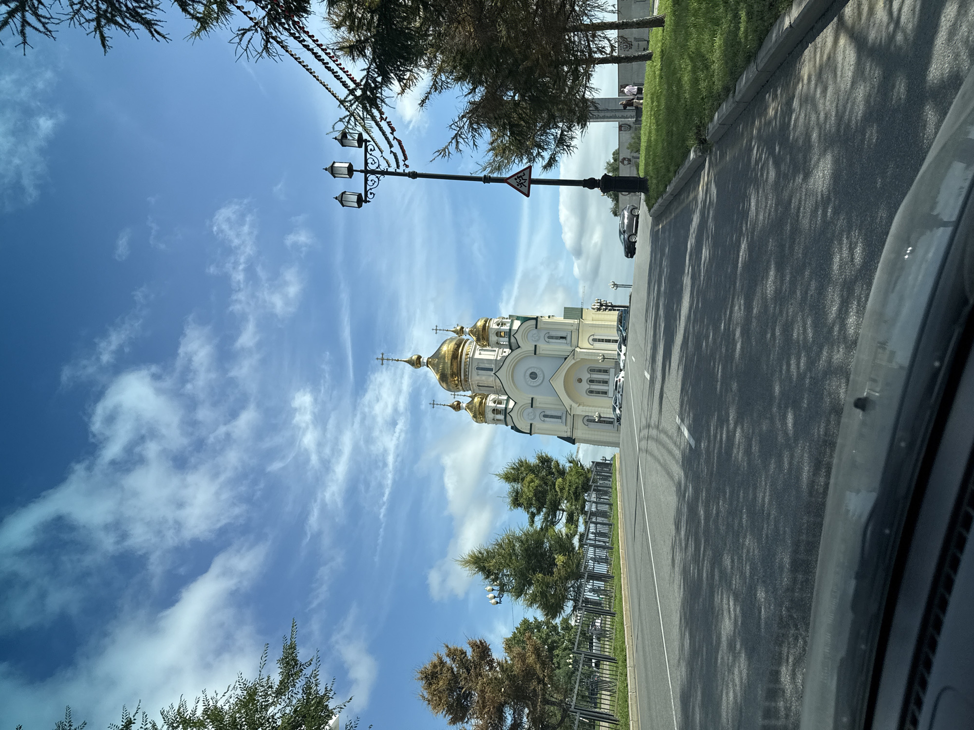 The image shows a view from a car looking towards a white Orthodox church with golden domes under a blue sky with white clouds. The church is located on a hill, and a paved road leads up to it. There are trees and a black metal fence along the side of the road. A lamppost with three lights stands on the right side of the road, along with a triangular traffic sign. The car's dashboard is visible in the foreground. The scene is likely in the city of Khabarovsk, Russia, given the architectural style of the church, which resembles the Transfiguration Cathedral in Khabarovsk. There are several cars parked in front of the church and two people walking on the grassy area to the right.