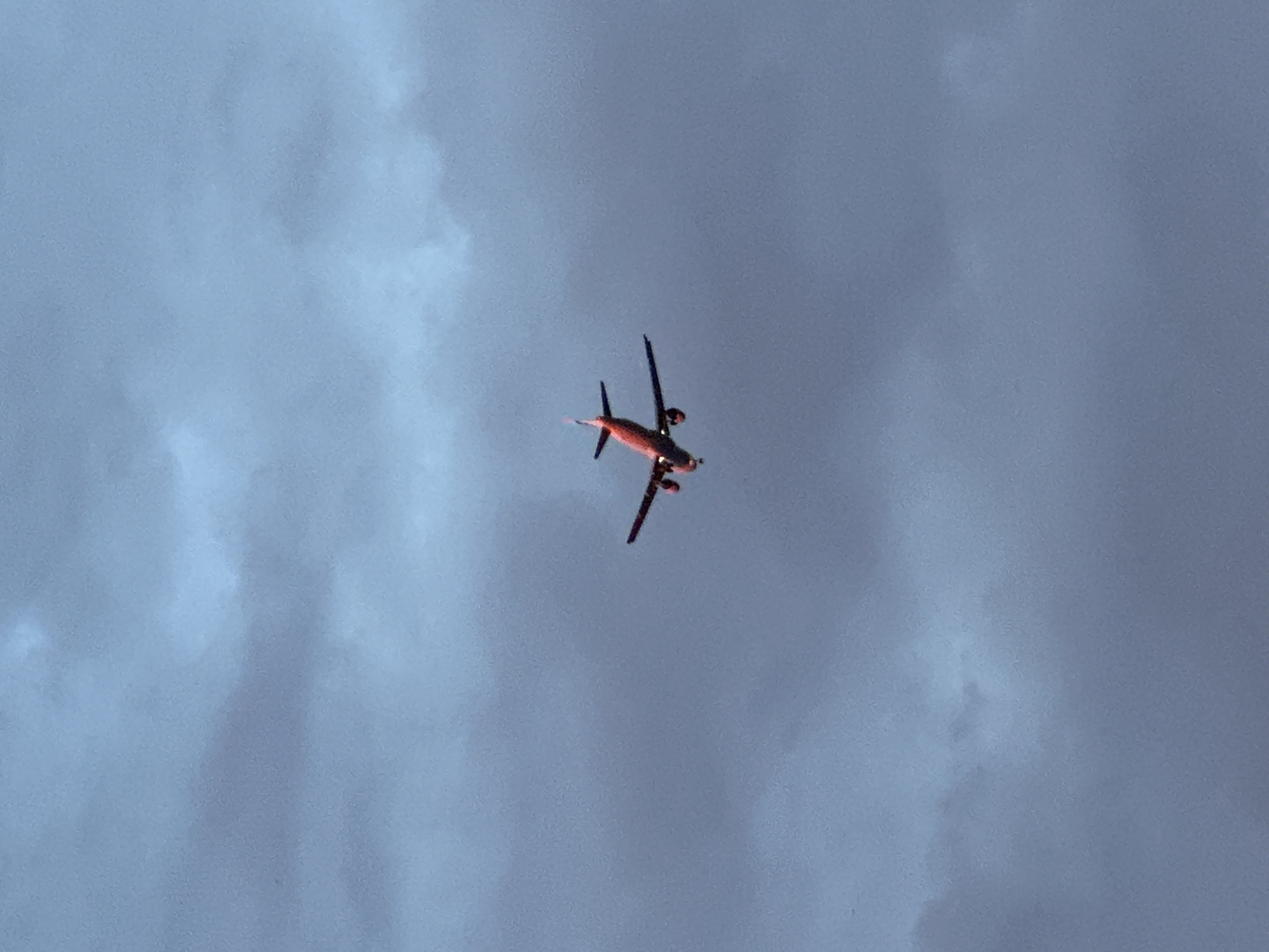 A low-angle shot captures a small propeller plane in flight against a cloudy, overcast sky. The plane is viewed from below and to the side, with its wings angled upwards. It appears to be a single-engine aircraft, possibly a seaplane, as suggested by the structure under its fuselage. The plane's body and wings are predominantly dark, with a reddish-orange glow emanating from its underside, possibly from the setting sun or internal lighting. The sky is a muted blue-gray, with soft, diffused clouds that create a slightly textured backdrop. There is no visible text in the image. The lighting suggests it could be late afternoon or early evening, given the glow on the plane and the subdued sky. The scene evokes a sense of travel or transit, with the plane as the sole subject against the vastness of the sky. The lack of clear landmarks makes it difficult to pinpoint a specific location, but the provided context indicates it is Tallinn, Estonia.