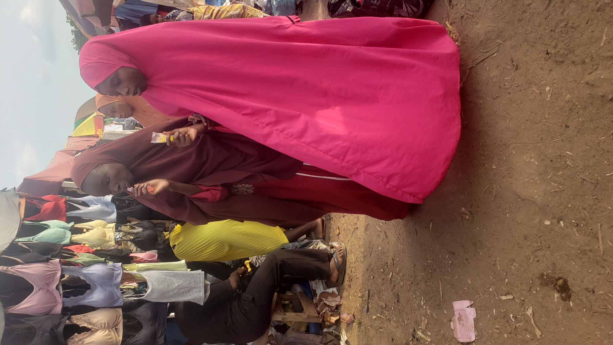 In Kakuri Gwari, Nigeria, an outdoor market scene unfolds, dominated by a stall displaying an array of bras of various colors and styles, hanging from a canopy. In the foreground, two young women, dressed in hijabs, stand close together. The woman on the left wears a maroon hijab and a matching dress, while holding a small, clear plastic container with a yellow substance and a pink item, seemingly a piece of candy or a snack. She is in the process of eating or preparing to eat it. To her right, a second woman is enveloped in a bright pink hijab and a matching dress. Her gaze is directed to her right, and she appears to be interacting with the woman beside her or looking at something off-camera. Behind them, a partially visible woman in an orange hijab stands, and further in the background, other individuals and market stalls are discernible. The ground is a dusty, uneven dirt surface, scattered with debris, including straw, small branches, and a discarded pink object, possibly a piece of packaging. To the left, a person wearing black clothing and sandals sits on a wooden stool, facing away from the camera, amidst a collection of bags and clothing items. The overall atmosphere suggests a bustling marketplace environment.