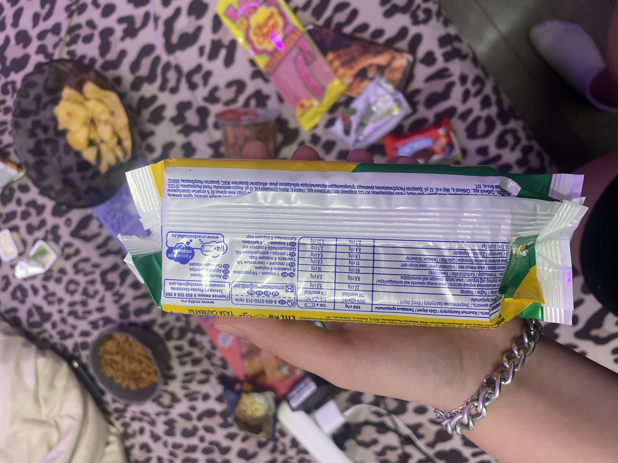 A person's hand with a silver bracelet is holding a food wrapper. The wrapper is green and yellow and has nutritional information printed on it in multiple languages. The background is a leopard-print fabric. There are several small packages of snacks scattered around the fabric. One of the snacks in the background appears to be a lollipop.