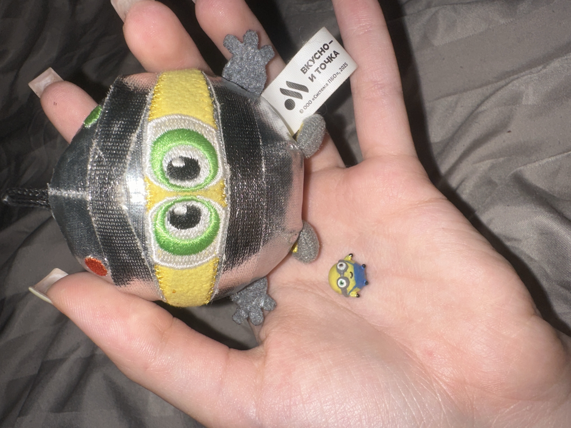 A close-up overhead shot captures a hand holding a collection of plush toys. The hand, which appears to be a woman's due to the long, light-colored artificial nails, is positioned against a textured gray fabric background. In the palm of the hand, two plush toys are visible. The larger toy, a silver and yellow plush character with large green eyes and small gray arms and legs, is held near the fingertips. Attached to its side is a white tag with black text and a logo. To the left and slightly below the larger toy, a much smaller, blue and yellow plush toy resembling a Minion character is placed in the palm. The overall lighting suggests an indoor setting. The image does not contain any elements that would allow for determining the location in Petropavlovsk-Kamchatsky, Russia.