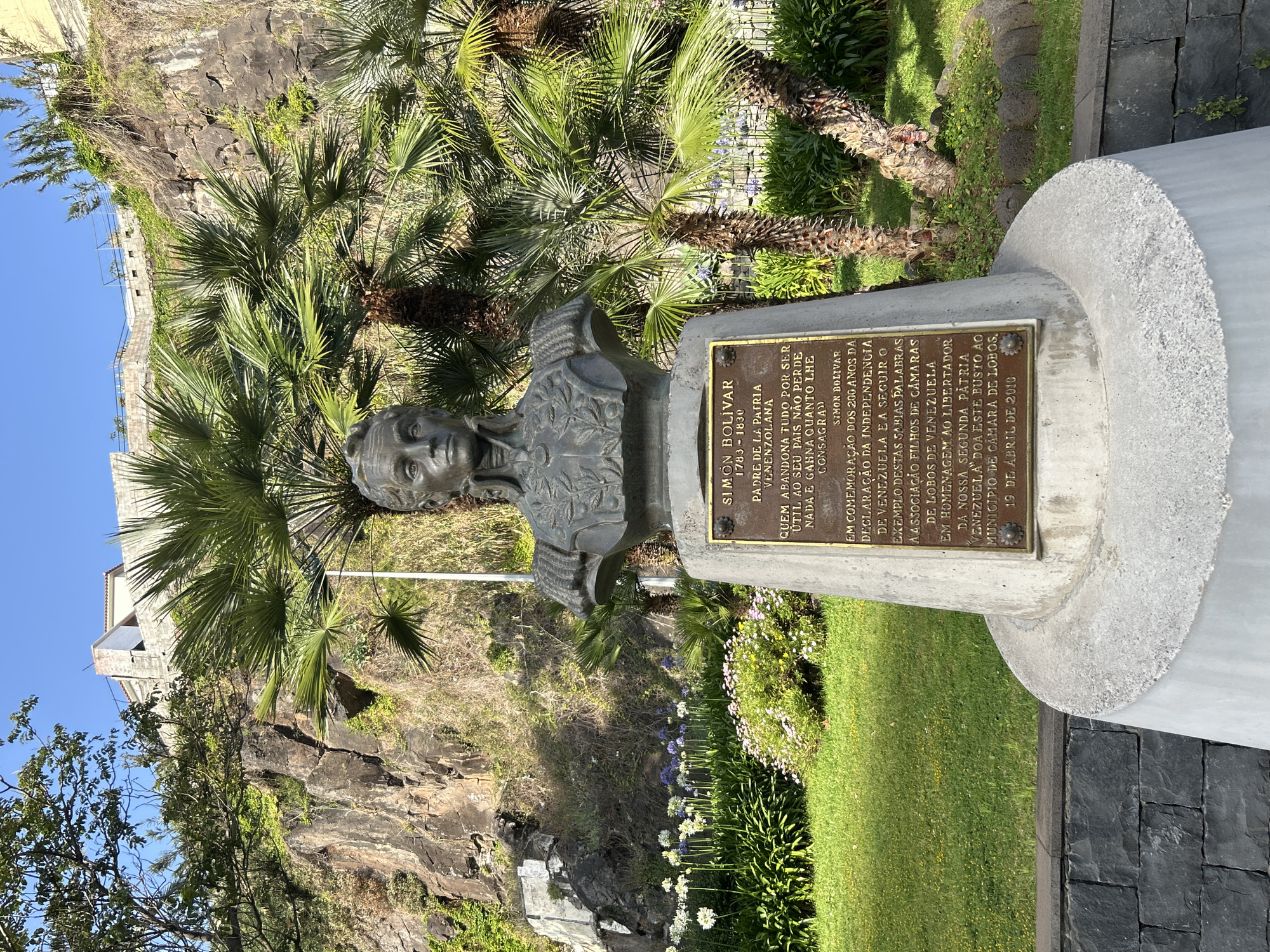 The image shows a bust of Simón Bolívar located in Câmara de Lobos, Portugal. The bronze bust sits atop a concrete pedestal. A plaque is mounted on the pedestal, displaying text in Portuguese, commemorating Bolívar and the 200th anniversary of Venezuela's independence. The plaque mentions the donation of the bust to the Municipality of Câmara de Lobos by the association of "Children of Câmara de Lobos of Venezuela." The date on the plaque is April 19, 2010. The monument is situated in a park or garden setting, evidenced by grass, flowers, and palm trees. A rocky hillside forms the backdrop. The weather appears sunny.