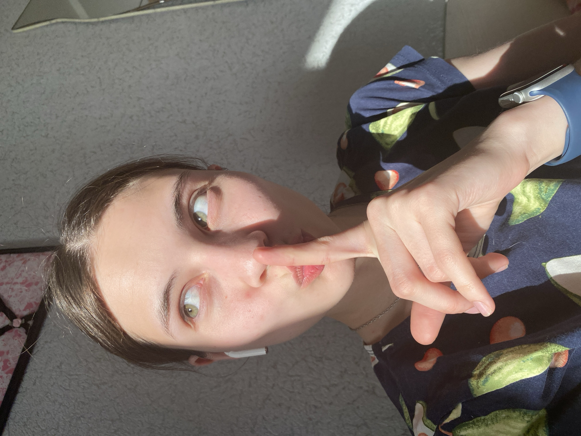 A young woman is facing the camera with her index finger touching her lips, possibly gesturing for silence. She has fair skin, light brown hair, and green eyes. She's wearing pajamas with a print that includes avocado and strawberry designs. On her left wrist, she is wearing a blue Apple Watch. She is indoors, and the photo is taken in bright, natural sunlight. The background consists of a gray carpet and part of a sofa or bed with a pink floral pattern.
This is not enough information to determine the location in Votkinsk, Russia.