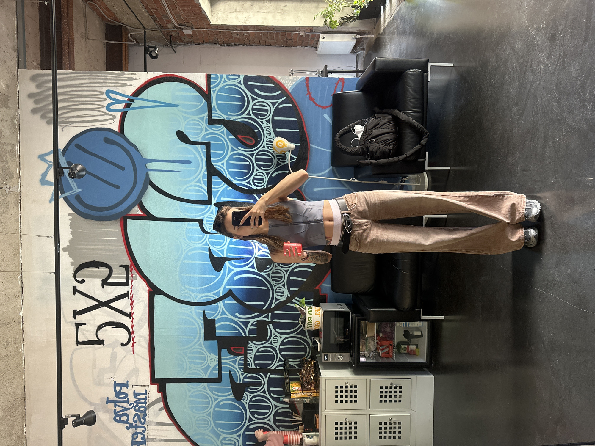 A young woman is taking a selfie in a loft-like space with exposed brick and concrete walls. She is wearing a grey crop top, brown pants, and sneakers. She is holding a red cup and her phone. In the background, there is a large graffiti mural with blue and white lettering. There is also a black leather couch, a small refrigerator stocked with drinks, a microwave, and some shelving units. A black bag is resting on the couch. The lighting suggests it is daytime. The overall mood is casual and urban. The graffiti on the wall appears to include the text "5XG" and some smaller, less legible text in Cyrillic.