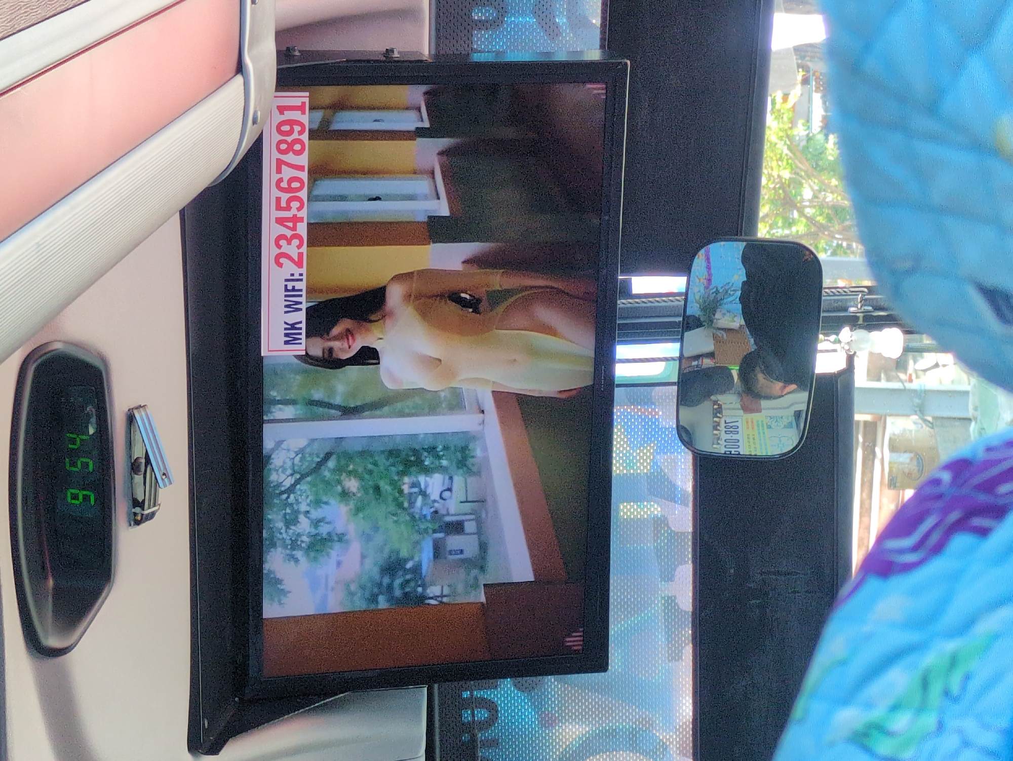 The image depicts the interior of a vehicle, possibly a bus, in South Quy Nhon Ward, Vietnam. A screen displays a scantily clad woman. A sign next to the screen reads "MK WIFI: 234567891" in red. A rearview mirror shows a reflection of the vehicle's interior. A digital clock on the wall reads "9:54". A seat with a pink and white cover is visible. Another seat, covered with blue patterned fabric, is partially visible in the lower right. The view through the vehicle's window shows trees and buildings.