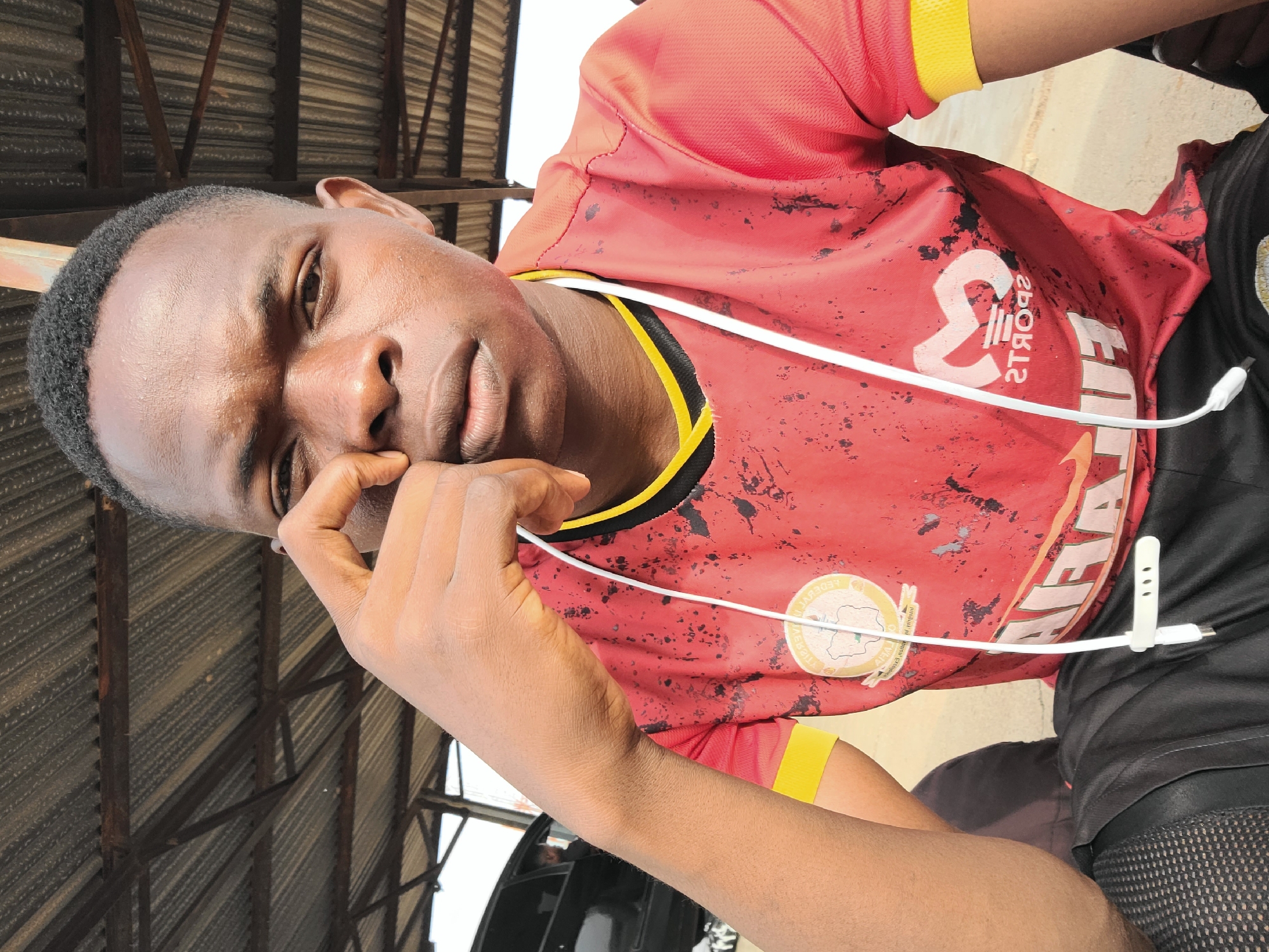 A young man is pictured from a high angle indoors under a corrugated metal ceiling, likely in Jos, Nigeria, judging from the crest on his red and yellow t-shirt and the person's features. He is looking at the camera, his fingers near his eye. He wears a red, short-sleeved athletic shirt with yellow trim, black shorts, and has a white cable clipped to his clothing. A blurry black object is in the lower left of the frame.