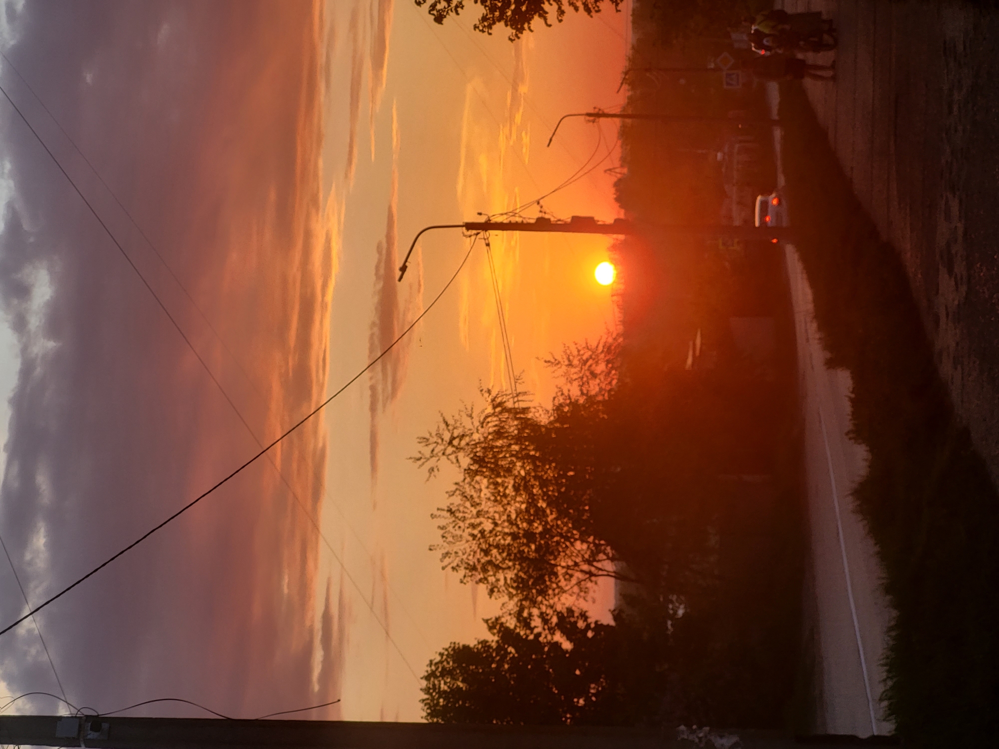 The image is a photograph of a sunset taken in a location presumed to be in the city of Chaikovsky, Russia. The sun is low in the sky, emitting a bright orange glow that washes across the scene. A road runs along the right side of the frame, with a white line marking its edge. Two people on a bicycle are barely visible riding on the shoulder of the road, along with a white car. Silhouetted trees and a utility pole with wires are present in the foreground and midground. The sky is a mix of orange, pink, and gray clouds. A large, black utility pole is in the lower left corner.