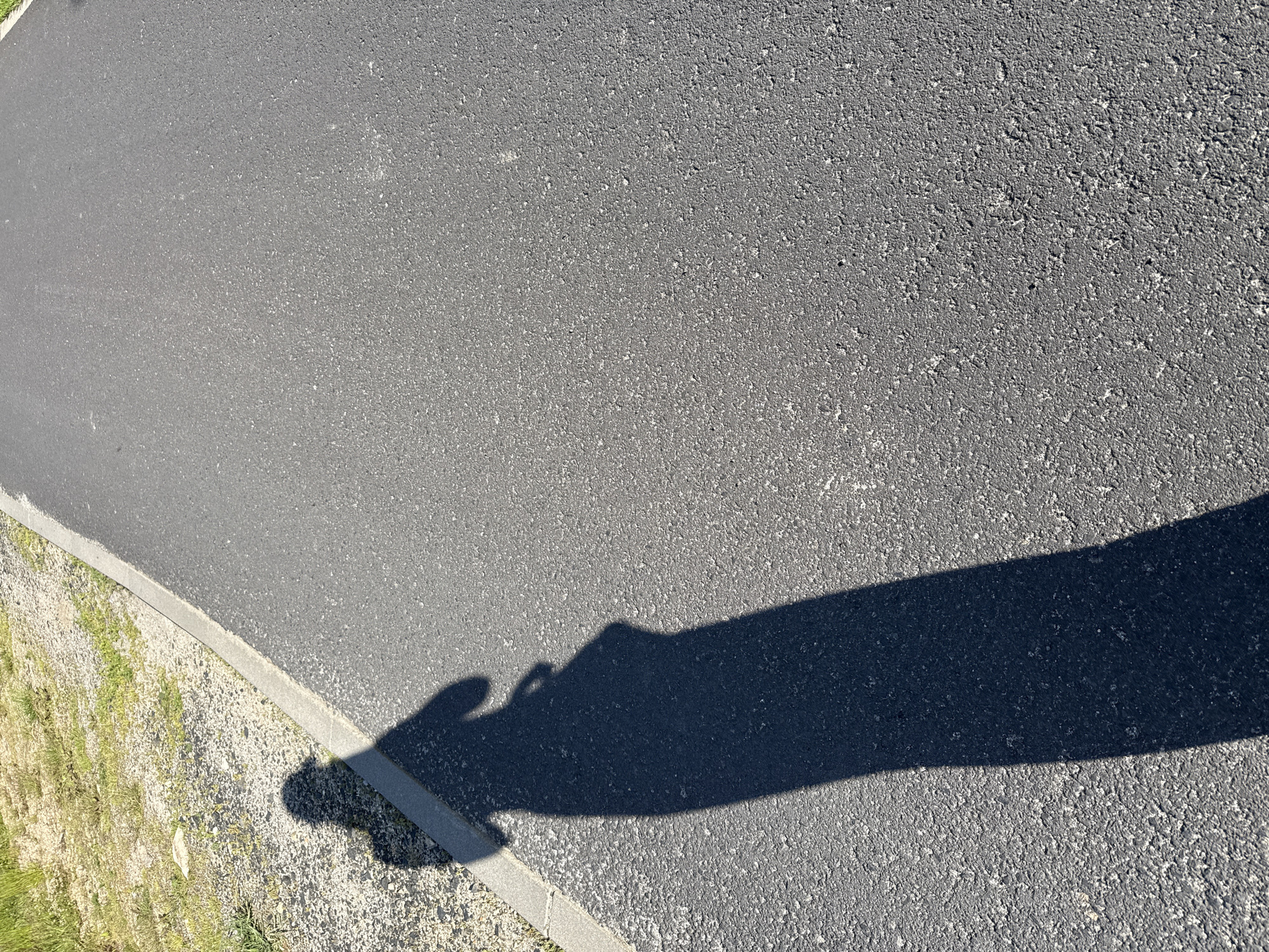 The image shows a shadow of a person casting on an asphalt path. The path is next to a grassy area with gravel. The shadow is long and indicates it is daytime. The location is identified as Szklarska Poręba, Poland. The scene is devoid of any other people or objects, focusing solely on the shadow and its surroundings.
