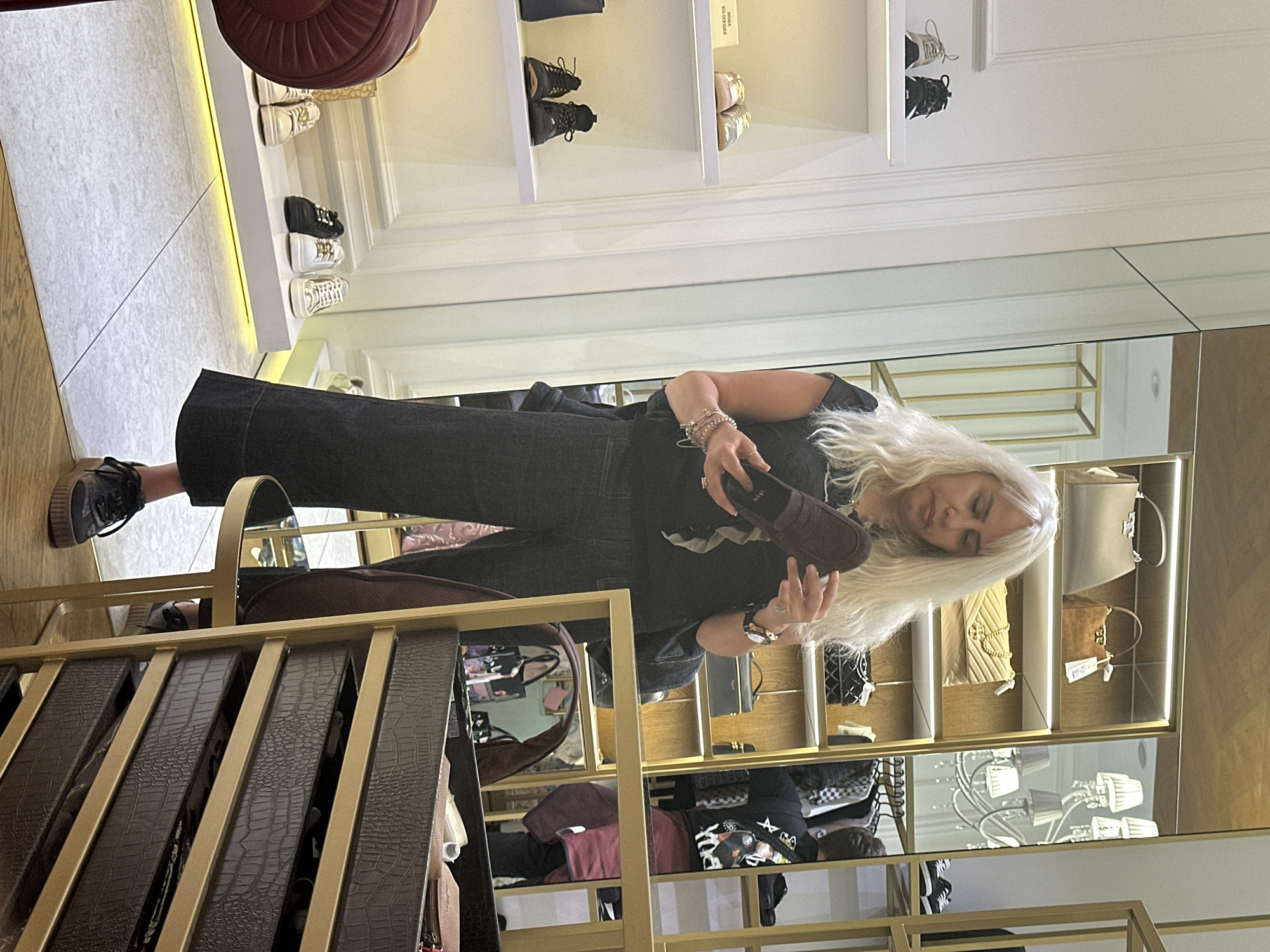 A woman with bleached blonde hair and wearing dark clothing is indoors, holding a brown shoe and looking at it. She is in a retail environment, possibly in Sofia, Bulgaria, where shelves of shoes and handbags are visible in the background. The foreground has a shelving unit with what appears to be leather belts with a crocodile pattern. The lighting is bright and even, suggesting a commercial setting.