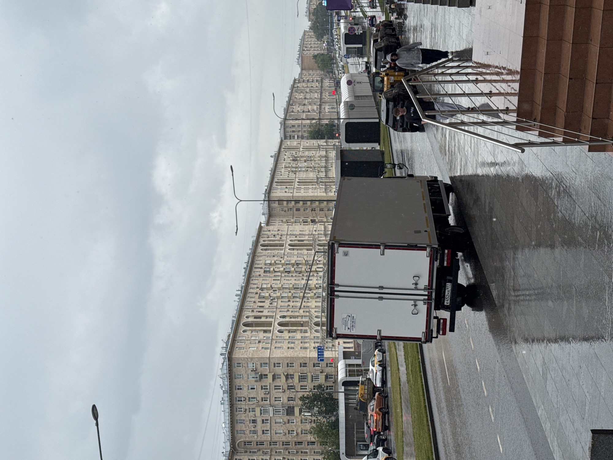 The image depicts a street scene in a city under overcast skies, with rain evident on the wet asphalt and pavement. A white box truck is prominently positioned in the foreground, facing away from the camera. To the left of the truck, a road is visible with several cars, including an orange SUV and a white and yellow taxi. Beyond the road, a long, multi-story building with numerous arched windows and balconies stretches across the background. Streetlights are visible along the road. In the mid-ground, several modern, minimalist structures with white curved roofs are lined up on the side of the road. Further down the street, more vehicles are visible, and on the right side of the image, two individuals are walking away from the camera on a wet sidewalk next to a set of brown tiled stairs with a metal railing. The overall atmosphere is somber and wet, characteristic of a rainy day.