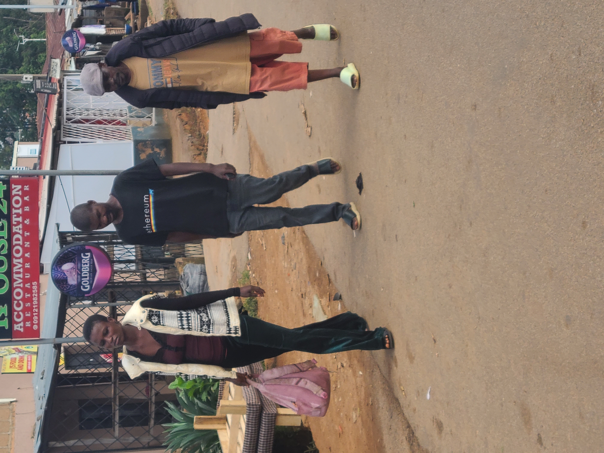 The scene captures three individuals walking along a paved road in what appears to be a street in Jos, Nigeria, as evidenced by the local signage. The first individual, located at the bottom of the frame, is a woman carrying a pink bag and a striped item. She wears a light-colored vest over a dark top and green trousers. Above her, a man sports a black t-shirt with the word "ethereum" printed on it, paired with dark jeans. At the top of the image, an older man wears a tan t-shirt with the word "TOMMY" visible. He is wearing orange shorts, a dark jacket, a baseball hat, and slippers.
The environment includes storefronts and buildings, notably "HOUSE 24 ACCOMMODATION RESTAURANT & BAR" sign. There's also signage for "GOLDBERG" (beer brand) visible. The road surface is relatively smooth.