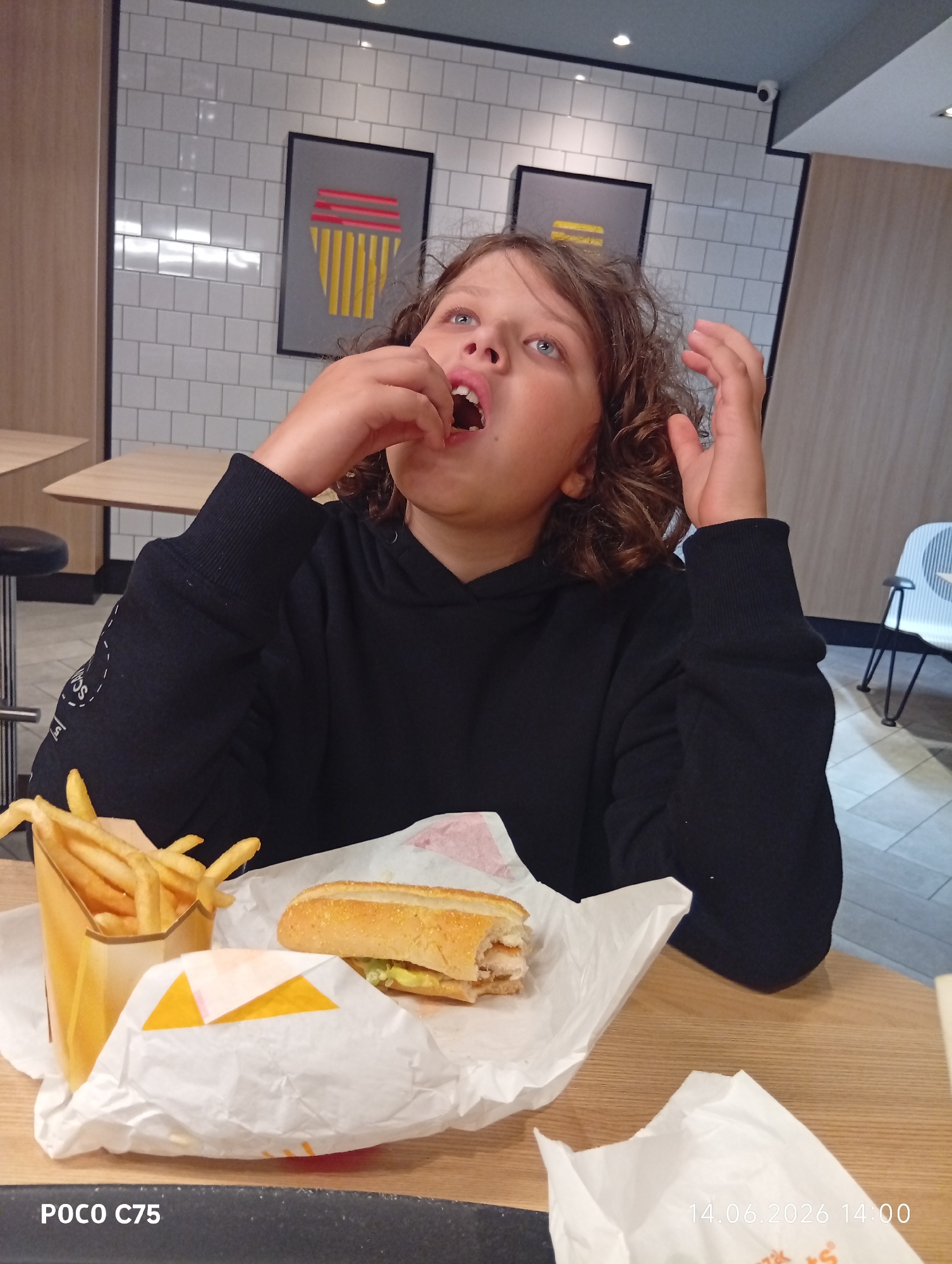 A young person with curly brown hair is eating a sandwich and french fries at a table in a brightly lit fast-food restaurant. The person is wearing a black hooded sweatshirt and is looking up with their mouth open as they place food into their mouth. In front of them are a container of french fries and a partially eaten sandwich. The walls of the restaurant are tiled in white, and there are framed pieces of art on the wall. The table appears to be made of wood. The date and time stamp on the image indicates it was taken on June 14, 2026, at 2:00 PM. The location is a fast-food restaurant in Wrocław, Poland.