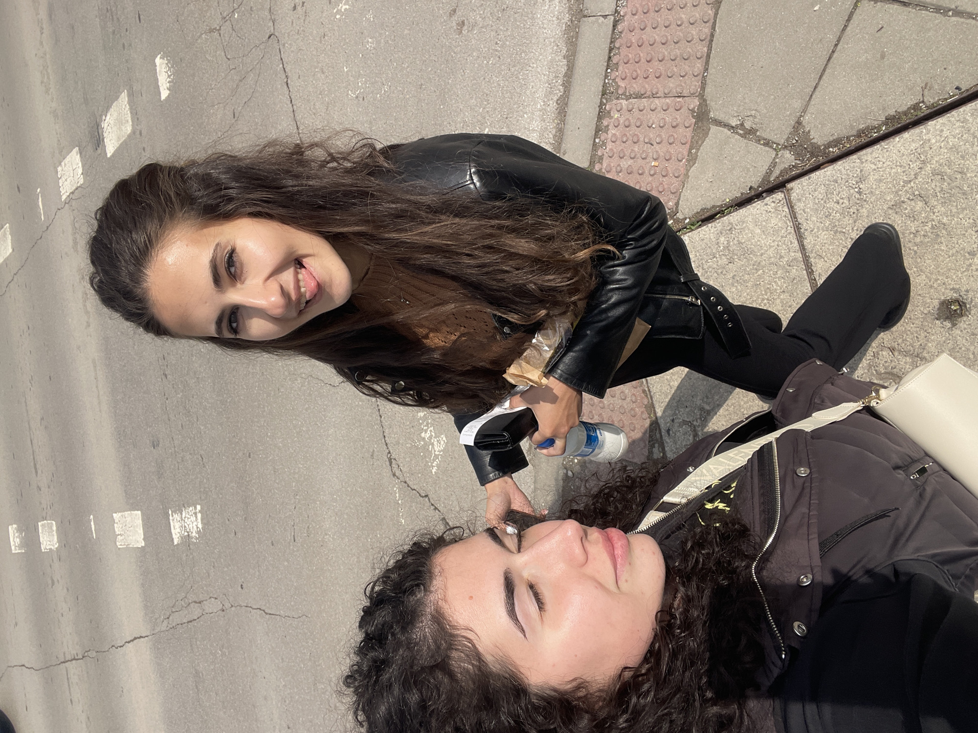 Two young women are outside in what appears to be the city of Sofia, Bulgaria. The woman on the left wears a black leather jacket and dark pants. She smiles, looking at the camera and sticking out her tongue slightly. She is holding a small bottle of water and a dark-colored object, possibly a phone. The other woman is lying on the ground, wearing a dark puffer jacket, with a white handbag beside her. The scene is lit by daylight, casting shadows on the pavement. The pavement has white markings. A textured edge suggests a sidewalk adjacent to the paved area.