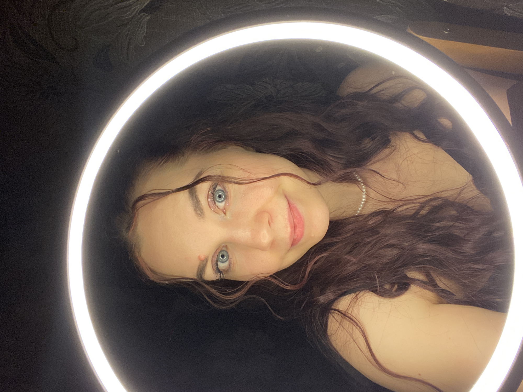 A woman with long, wavy brown hair is posing for a selfie in front of a ring light. She is wearing a pearl necklace and appears to be smiling slightly. Her eyes are blue. The background is dark and indistinct. There's no information in the image to confirm that the location is Chaikovsky, Russia.