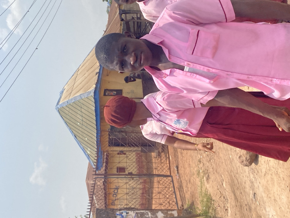 This image depicts an outdoor scene in Afikpo, Nigeria, under a bright, sunny sky with scattered clouds. In the immediate foreground, a young dark-skinned male, appearing to be a student, is positioned on the right, looking directly at the viewer with a neutral expression. He wears an unbuttoned light pink short-sleeved collared shirt over a white undershirt. To his left and slightly behind him, a young dark-skinned female, also appearing to be a student, is captured in motion, her back largely facing the camera but her head turned slightly to the left. She wears a dark red beret, a light pink short-sleeved collared shirt featuring a school emblem on the left chest, and a dark red long skirt. Her left arm is bent, revealing a thin light-colored bracelet. A third individual, partially visible on the far right, also wears a pink shirt.
The ground beneath the subjects is an unpaved, reddish-brown dirt path. In the midground, a prominent yellow single-story building with a distinctive pitched roof is visible. The roof appears to be constructed of corrugated metal, varying in color from light tan to olive, with a blue trim along its eaves. A dark metal gate with intricate scrollwork, behind which the word "BLOCK" is written in white on a wall, stands to the left of the main yellow building. Another blurred, dark-skinned figure is observed leaning or bending near the central yellow building to the right. Utility lines stretch horizontally across the upper portion of the frame against the sky.