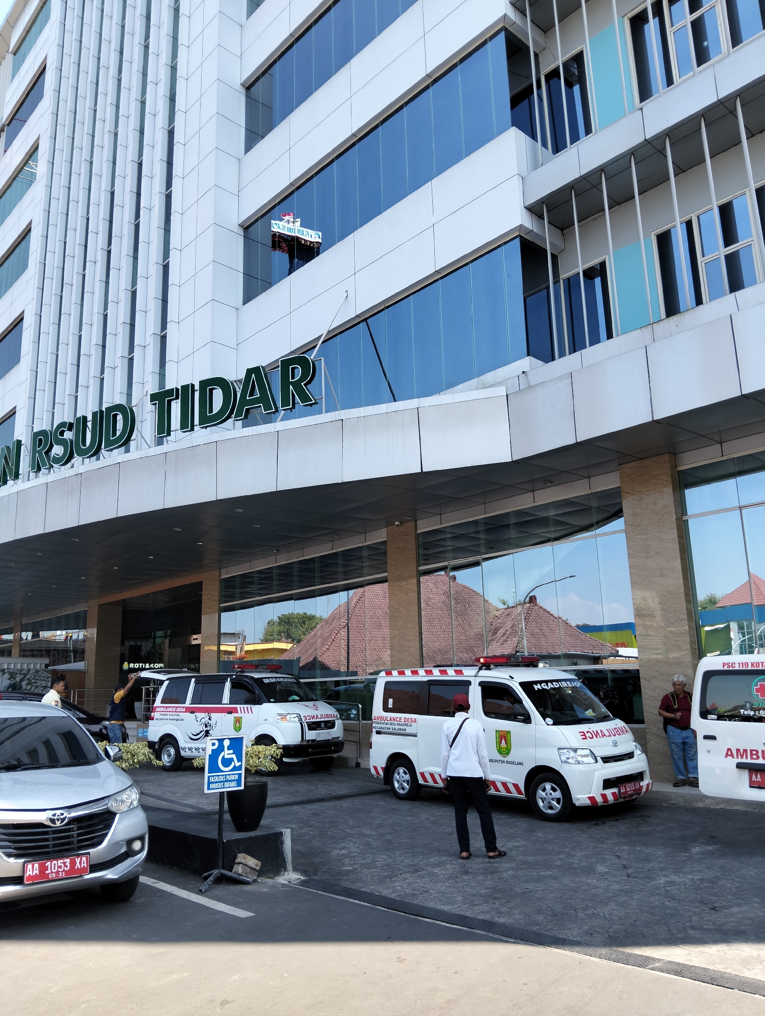 The image shows the exterior of a modern hospital building in Magelang, Indonesia, identified by the sign "N RSUD TIDAR". The building features a white facade with reflective blue windows. In front of the hospital, several vehicles are parked. A silver Toyota Avanza is in the foreground on the left, with license plate "AA 1053 XA". Behind it, two white ambulances are parked. The ambulance on the left has markings including "AMBULANCE DESA" and "KECAMATAN BANDONGAN", and "ABUPATEN MAGELANG". The ambulance on the right has "PSC 119 KOTA" visible. There is a disabled parking sign with a wheelchair icon and Indonesian text. Two men are standing near the ambulances, and one man is standing near the silver car. The overall environment appears to be a bright, sunny day with the hospital entrance and parking area visible.