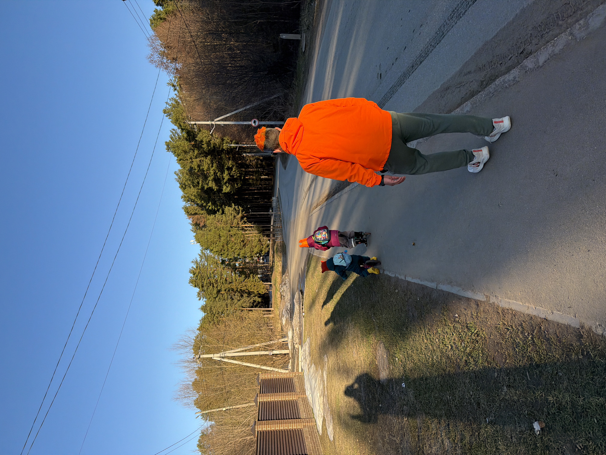 The image depicts a scene on a road in what is likely a residential area. In the foreground, an adult wearing an orange jacket and cap, and green pants, is walking on the asphalt road surface. Further down the road and slightly behind, two children are present. One is wearing a purple jacket and the other a blue jacket. The road is bordered by a grassy area. A fence and trees are visible in the background. Power lines stretch overhead against a clear blue sky. Considering the location's mention of Chaikovsky, Russia, the environment suggests a semi-urban setting with a blend of residential and natural elements. The road shows signs of vehicle traffic.