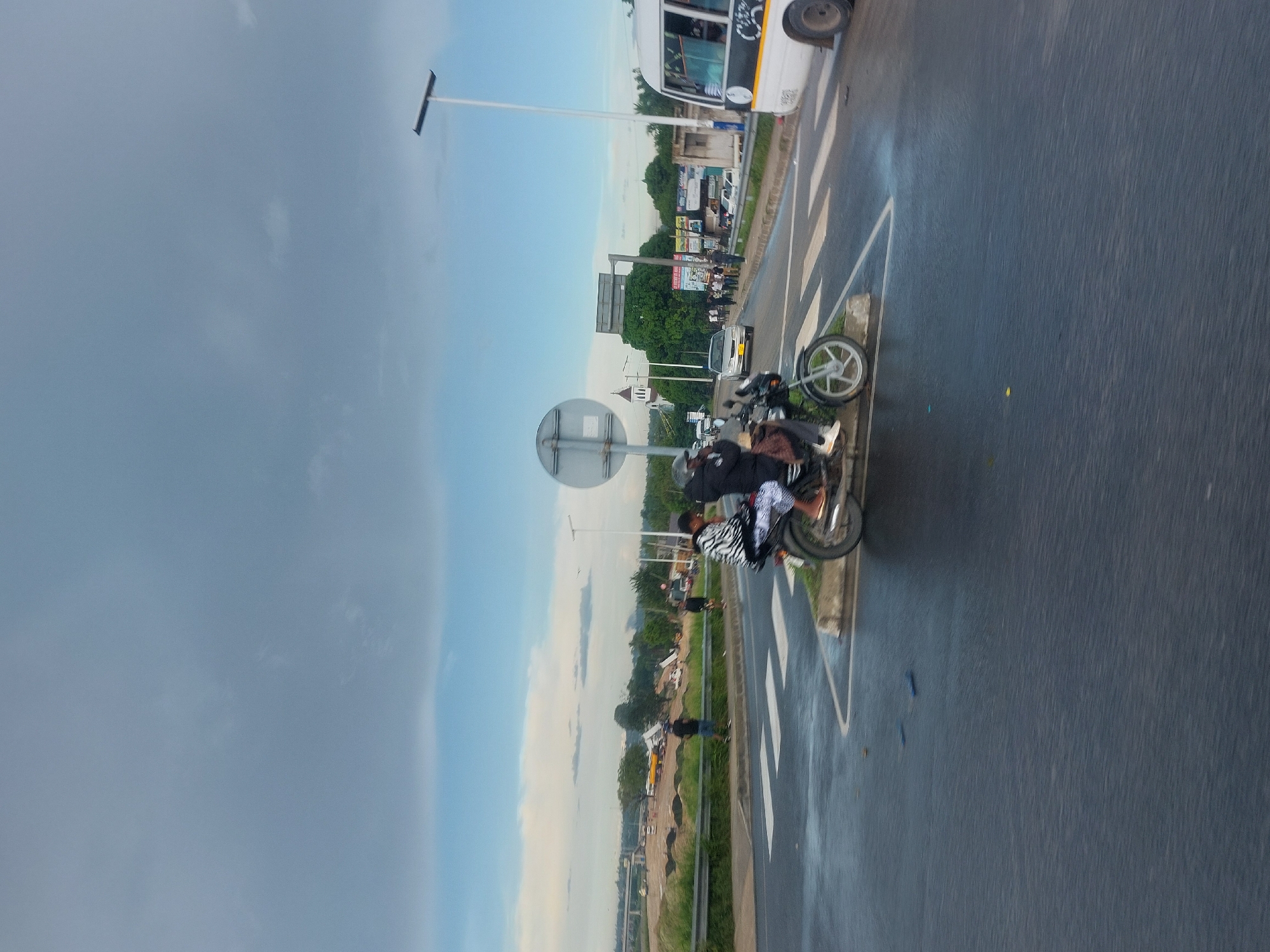 The image presents a scene on a roadside in what appears to be a tropical environment. Two people are on a motorcycle. The road is paved with white lane markings. A large bus is visible on the road, along with other vehicles in the distance. There are trees and structures present in the background. The sky is partly cloudy. The image is possibly in Dar es Salaam, Tanzania.