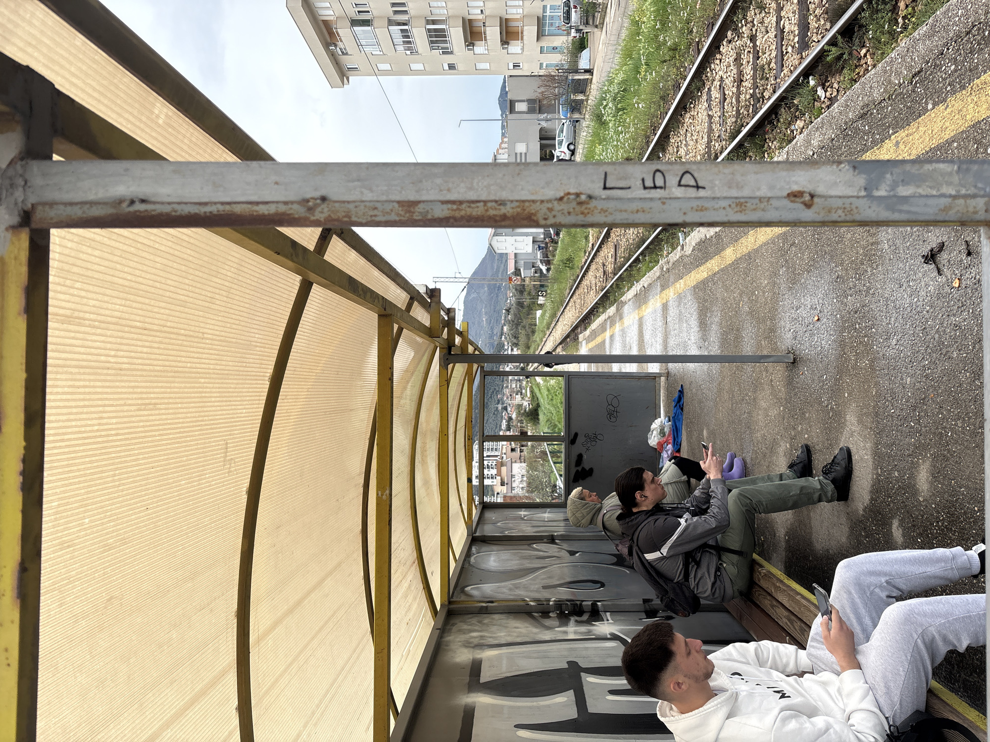 The image depicts the train station in Bar, Montenegro. Three individuals are sitting on a bench in a covered waiting area. Two are looking at their phones. One wears a white hoodie, the second is in a grey jacket and green pants. A third person is sitting at the end of the bench, also appearing to use a phone. The waiting area has graffiti on the walls. Outside the waiting area, train tracks run parallel to the platform. Buildings are visible in the background, with mountains in the distance. The sky is overcast. The overall impression is of a somewhat dilapidated train station.