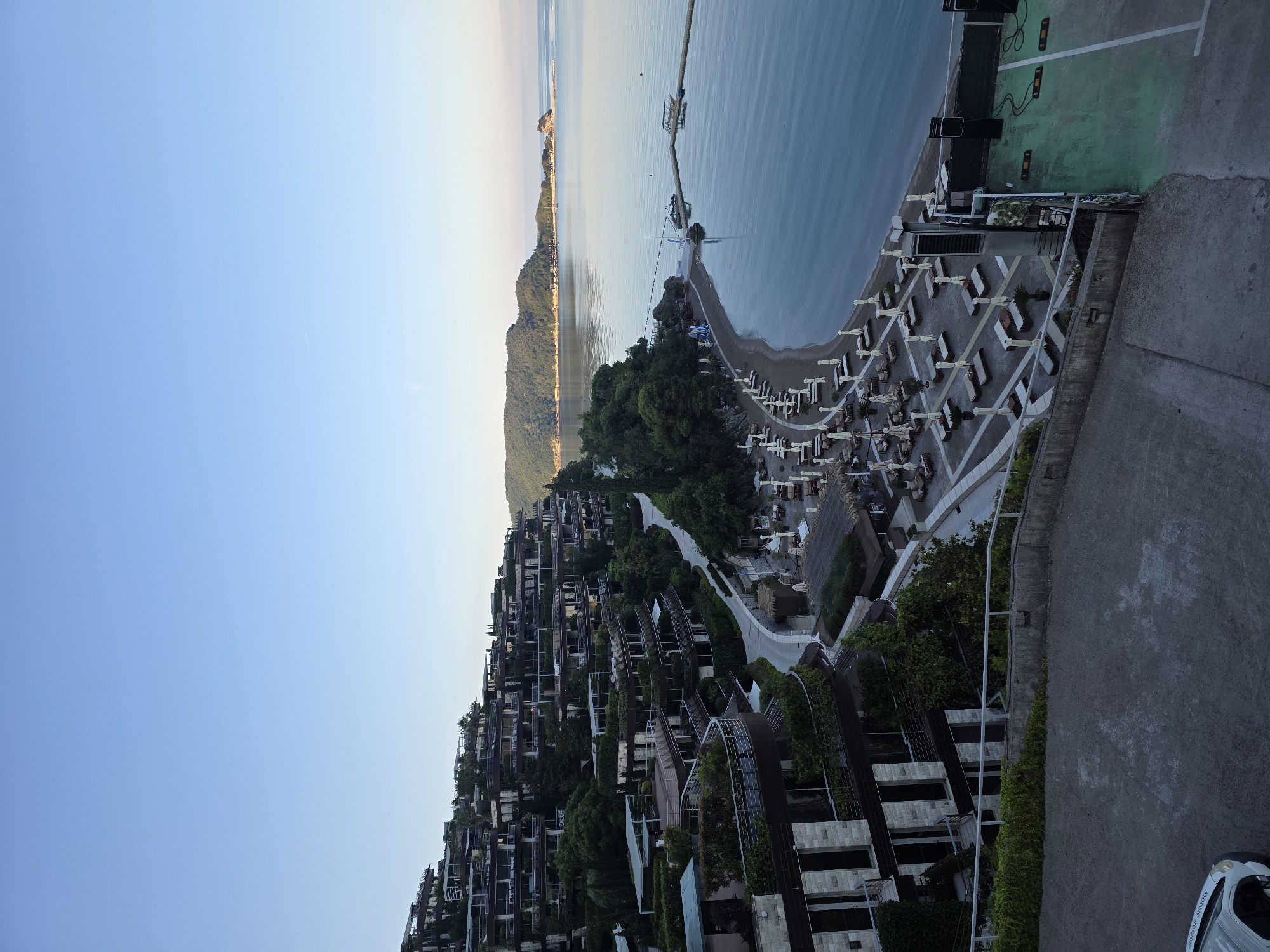 This is an elevated, wide-angle, medium-shot, outdoor, vertical photograph taken during the daytime in Budva, Montenegro. The scene captures a resort complex cascading down a hillside towards a beach and the Adriatic Sea.
In the foreground, a paved surface with some vegetation is visible. A portion of a white vehicle is parked on this surface on the lower left.
The mid-ground features the resort's multi-level architecture, characterized by terraced buildings with balconies and abundant greenery, including trees and shrubs integrated into the structures. A curved pathway winds through the complex. Below the resort, a sandy beach is set up with rows of lounge chairs and umbrellas, some of which are white and others are brown. Adjacent to the beach is a dark-colored, partially covered structure.
The background showcases the tranquil, calm, blue water of the Adriatic Sea. A long, narrow pier extends from the shore into the water, with at least two small boats docked. In the distance, lush green hills form the coastline, and a small island or rock formation is visible in the sea. The sky is clear and a pale blue, indicating it might be early morning or late afternoon.
No people are prominently featured or engaged in any discernible actions within the frame. The environment suggests a peaceful, resort-like setting. The overall lighting is soft, consistent with natural daylight, and there are no harsh shadows.