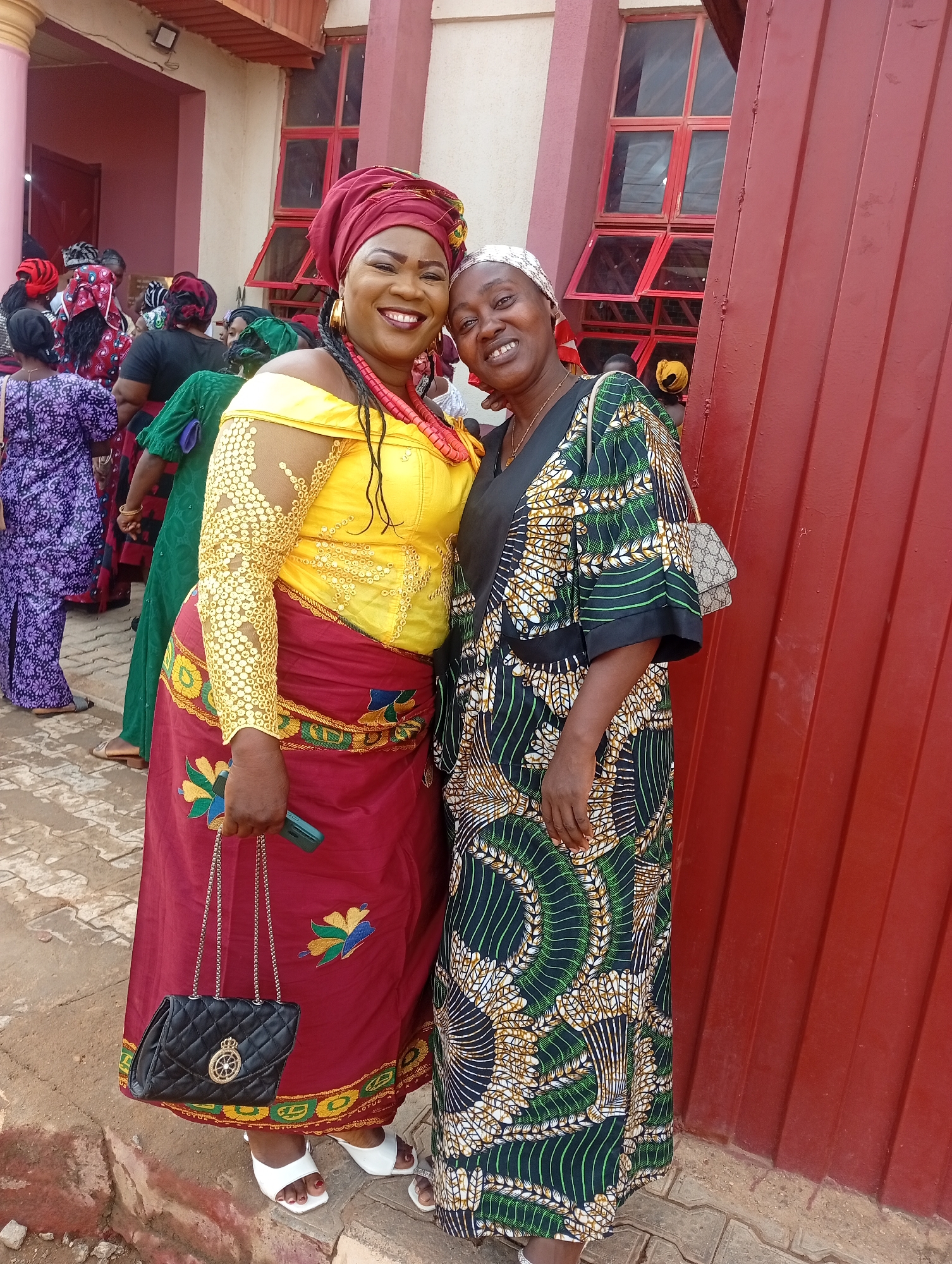 Two women stand closely together outside a building with a red metal wall in Narayi, Nigeria. The woman on the left wears a maroon head wrap, a yellow long-sleeved dress, and a maroon skirt. She is carrying a black purse and holding a smartphone. The woman on the right wears a patterned green and brown dress and a white head wrap. Other people in traditional clothing are visible in the background. The building is pink and red, with red-framed windows. The ground is paved with interlocking concrete pavers.