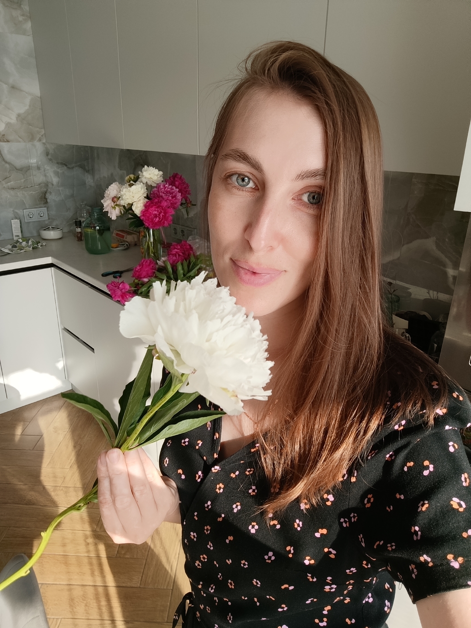 A woman with long, brown hair and blue eyes smiles directly at the camera, holding a large white peony in her right hand. She is wearing a black dress with a scattered pattern of small pink and orange flowers. In the background, a kitchen counter is visible with a vase of pink and white peonies and a green glass pitcher filled with a light-colored liquid. The kitchen has white cabinets and gray marble-patterned backsplash. Sunlight streams into the room, casting shadows on the wooden floor. The scene appears to be indoors, likely in a home kitchen in Chaikovsky, Russia. The overall mood is bright and cheerful, suggesting a pleasant day. There is no visible text in the image.
