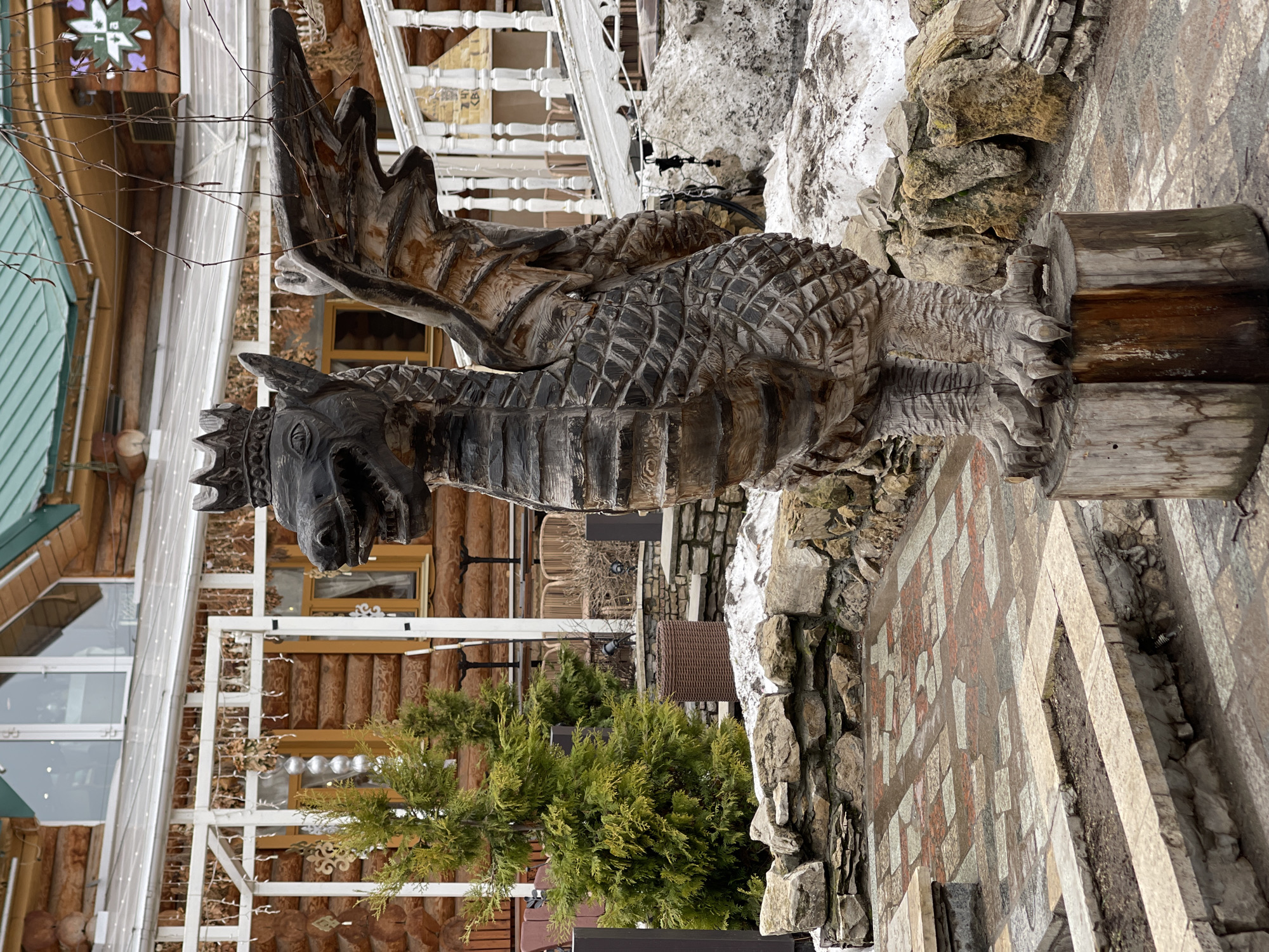 The image shows a wooden statue of a dragon in Kazan, Russia. The statue is detailed, with scales, wings, and a crown. It stands on a wooden post surrounded by rocks. In the background, there is a building with log walls and white trim, possibly a restaurant or tourist attraction. A small green tree sits in front of the building, near the statue. The ground is covered with patterned stone tiles. Patches of snow are visible near the statue.