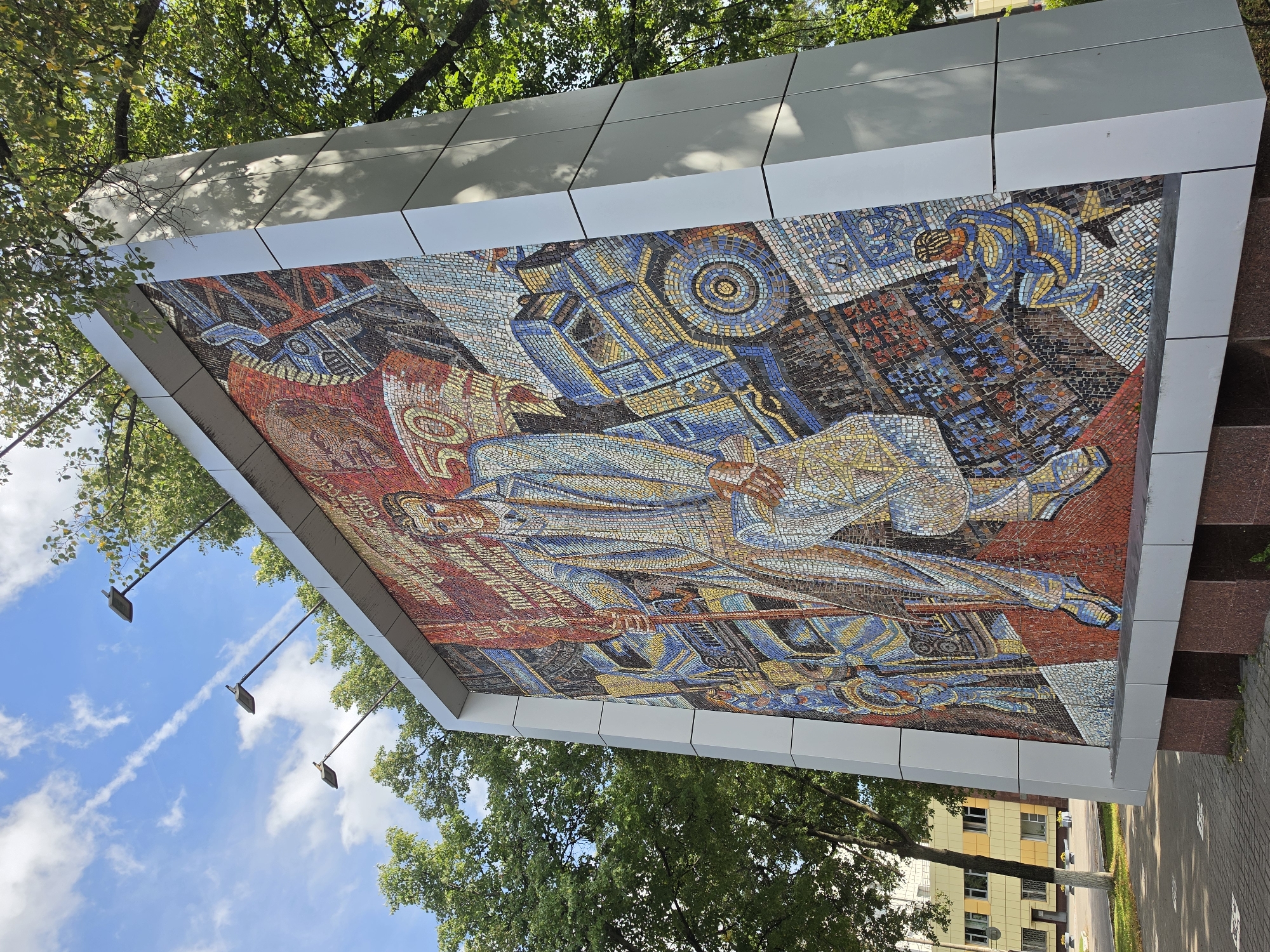 The image shows a large mosaic artwork situated outdoors in a city setting, likely in Nizhny Novgorod, Russia, given the context of similar mosaic artworks found in the region. The mosaic is mounted on a tall, modern, grey metal-clad structure with angular supports. The artwork itself is vibrant and detailed, depicting a central figure of a man in a suit holding a banner, surrounded by scenes of industry and progress. There are representations of trucks, machinery, and smaller figures engaged in various activities. Text in Cyrillic script is visible within the mosaic, suggesting it commemorates an event or achievement. The mosaic is illuminated by the sun, casting shadows from the surrounding trees. In the background, a building with yellow and white facades can be seen, along with a paved area and sidewalk. The sky above is blue with scattered white clouds and contrails from an aircraft. Several streetlights are mounted on poles extending from the top of the mosaic structure. The overall scene conveys a sense of public art installation in an urban environment.