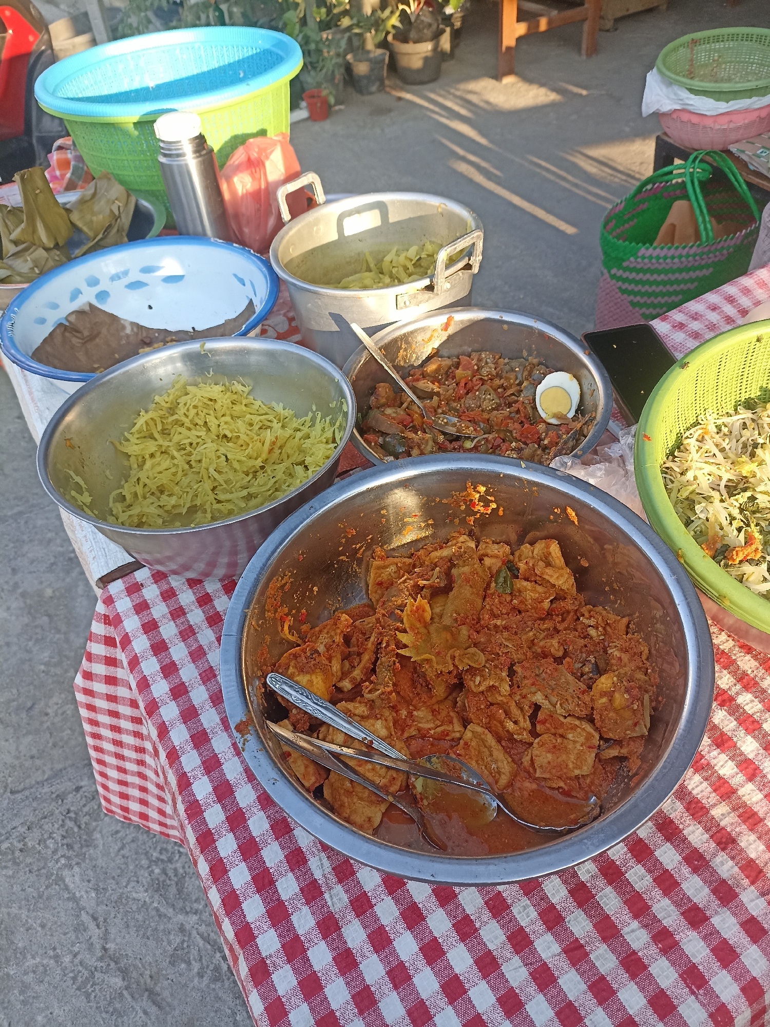 This image depicts an outdoor food stall in Mantup, Indonesia, on a sunny day. A red and white checkered tablecloth covers the surface of the stall, on which several bowls and containers of food are arranged. In the foreground, a large metal bowl contains a reddish-brown dish with chunks of meat and a saucy texture. Tongs are placed inside this bowl. To its right, a green plastic basket holds a salad-like mixture of shredded vegetables. Behind these, a smaller metal bowl contains a yellow, shredded food item. Next to it, another metal bowl holds a reddish dish with visible pieces of eggplant or similar vegetables, and a hard-boiled egg half. Further back, a metal thermos is placed next to a red plastic bag. To the left of the thermos, a blue and green plastic basin contains what appears to be wrapped food items. In the background, more food containers and green plastic baskets are visible, along with plants in pots and a wooden structure, suggesting the stall is set up in a yard or open space. The ground is concrete, and the sunlight casts shadows on it, indicating it is late afternoon.